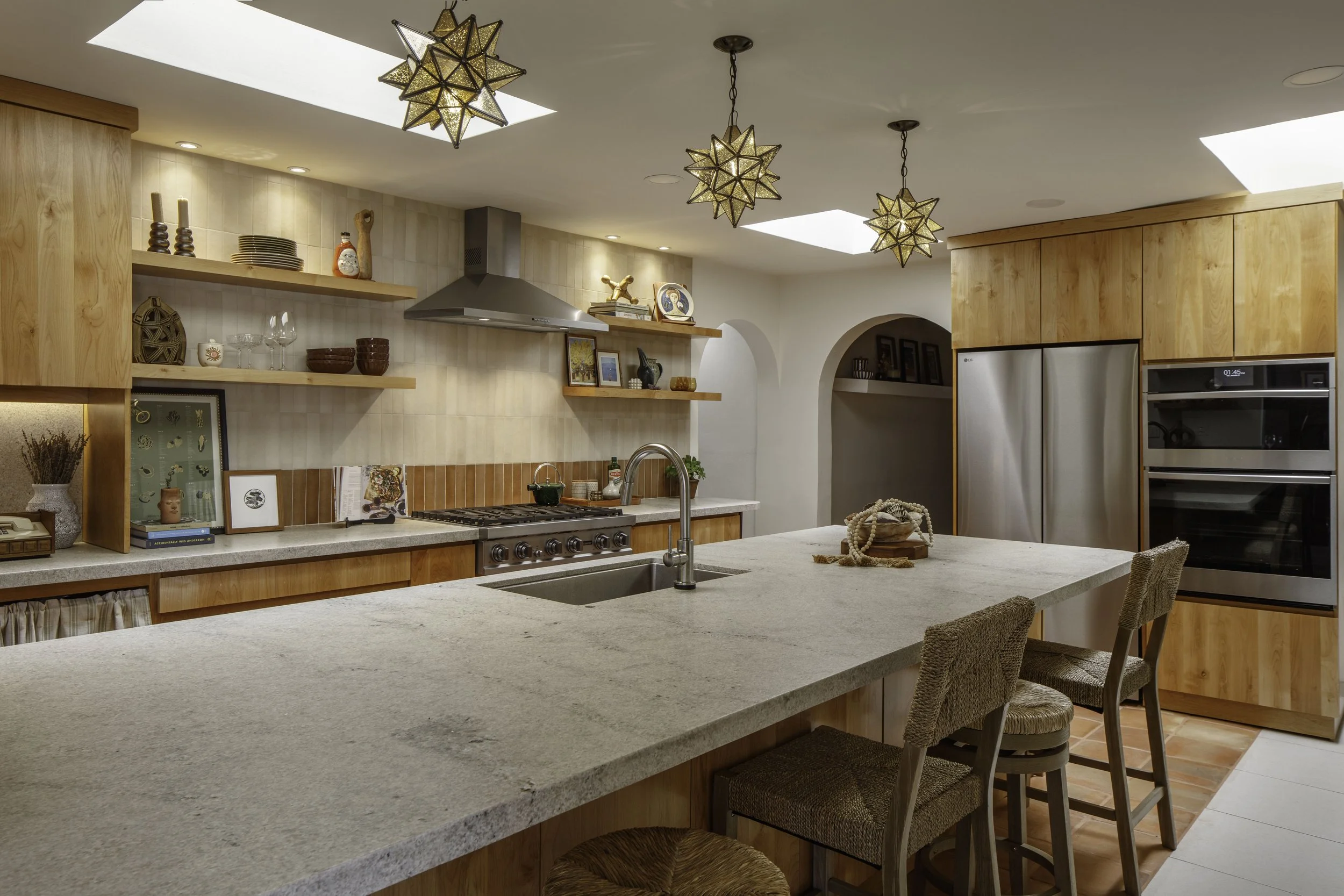 Modern kitchen with light wood cabinets, open shelves, stainless steel appliances, a large center island with stools, decorative star-shaped pendant lights, and skylights.