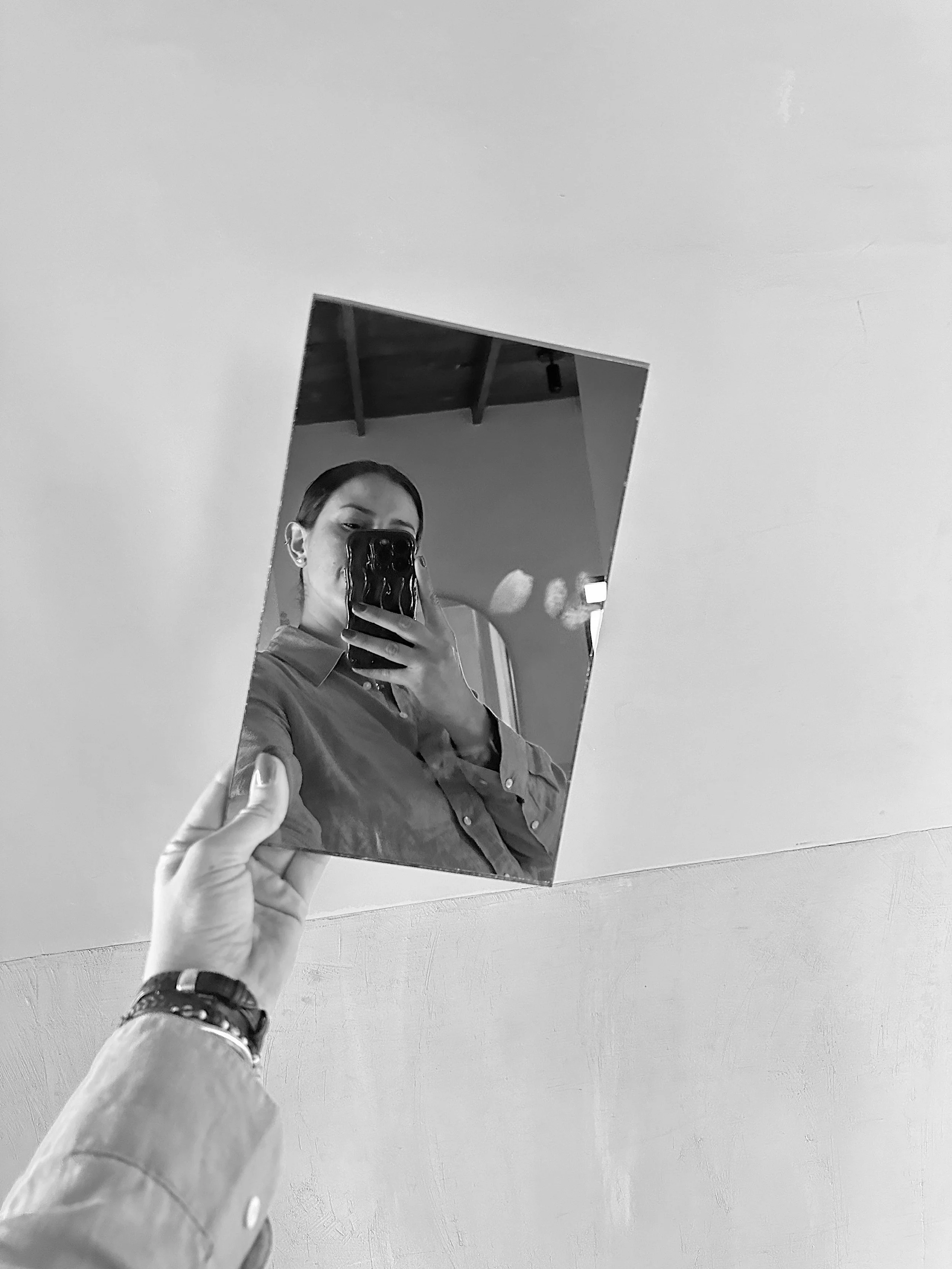 Person holding a small mirror reflecting their own image on a textured wall background in black and white.