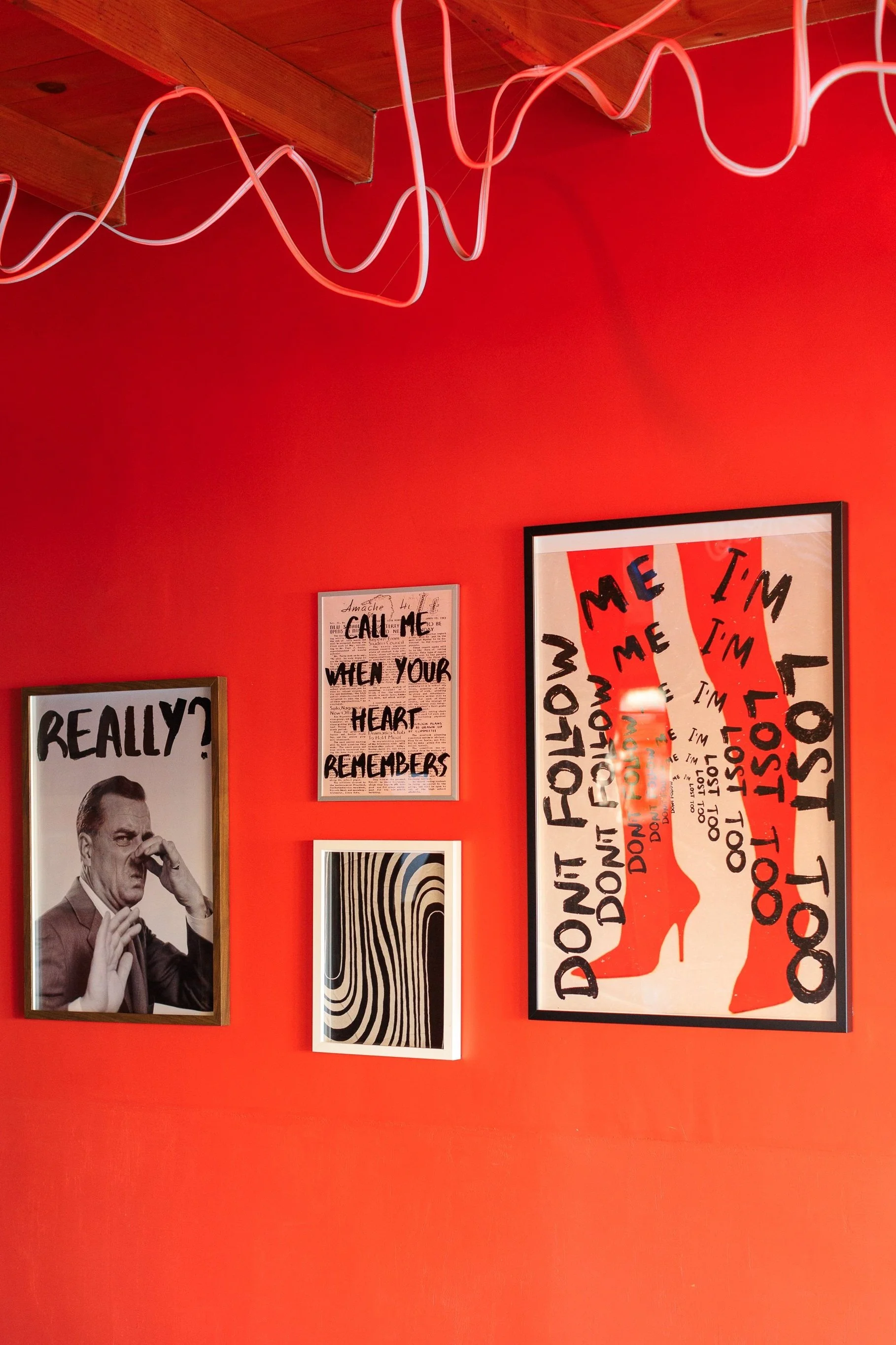Red wall with various framed artworks, including a black and white photo of a man with text "REALLY?", abstract black and white stripes, text art with "CALL ME WHEN YOUR HEART REMEMBERS", and a piece with red high heels and text "DON'T FOLLOW ME I'M 