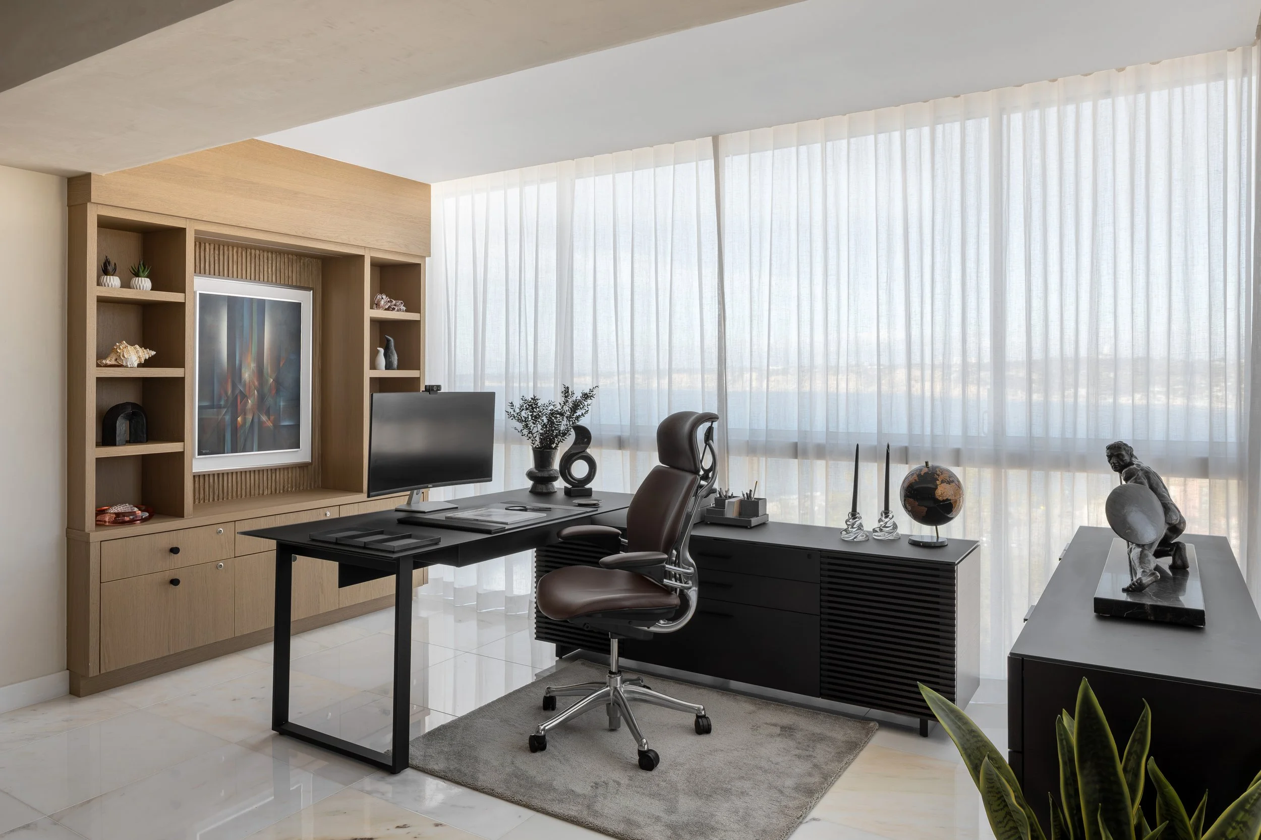 Modern home office with a large desk and ergonomic chair, featuring a computer monitor, artistic decor, and a globe, set against floor-to-ceiling windows with sheer curtains. The room includes built-in shelves with decorative items and a framed abstr