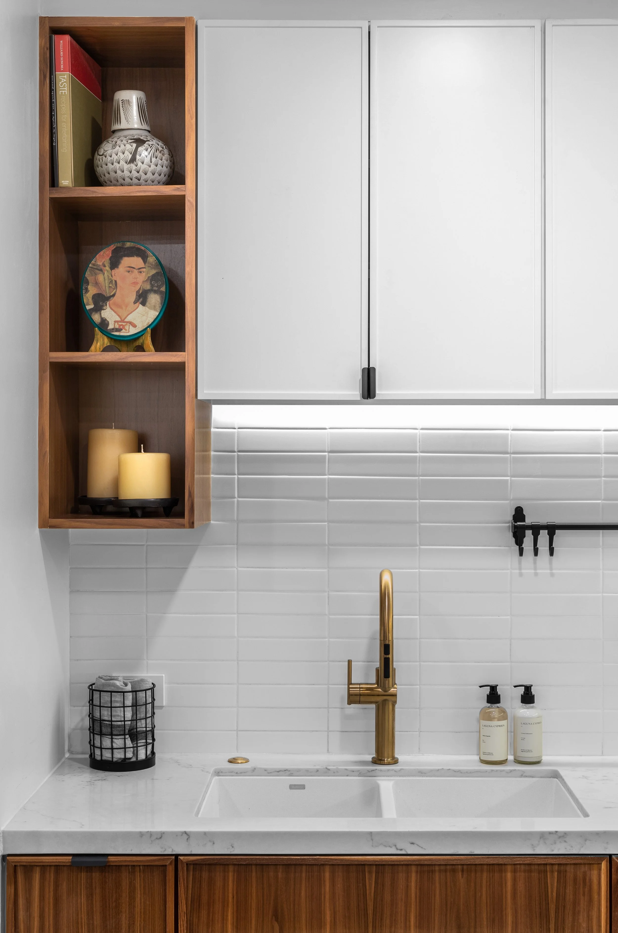 Modern kitchen corner with a white tile backsplash, wooden shelving, and white cabinets. Features a gold faucet, soap dispensers, and neatly placed books, candles, and decorative items.