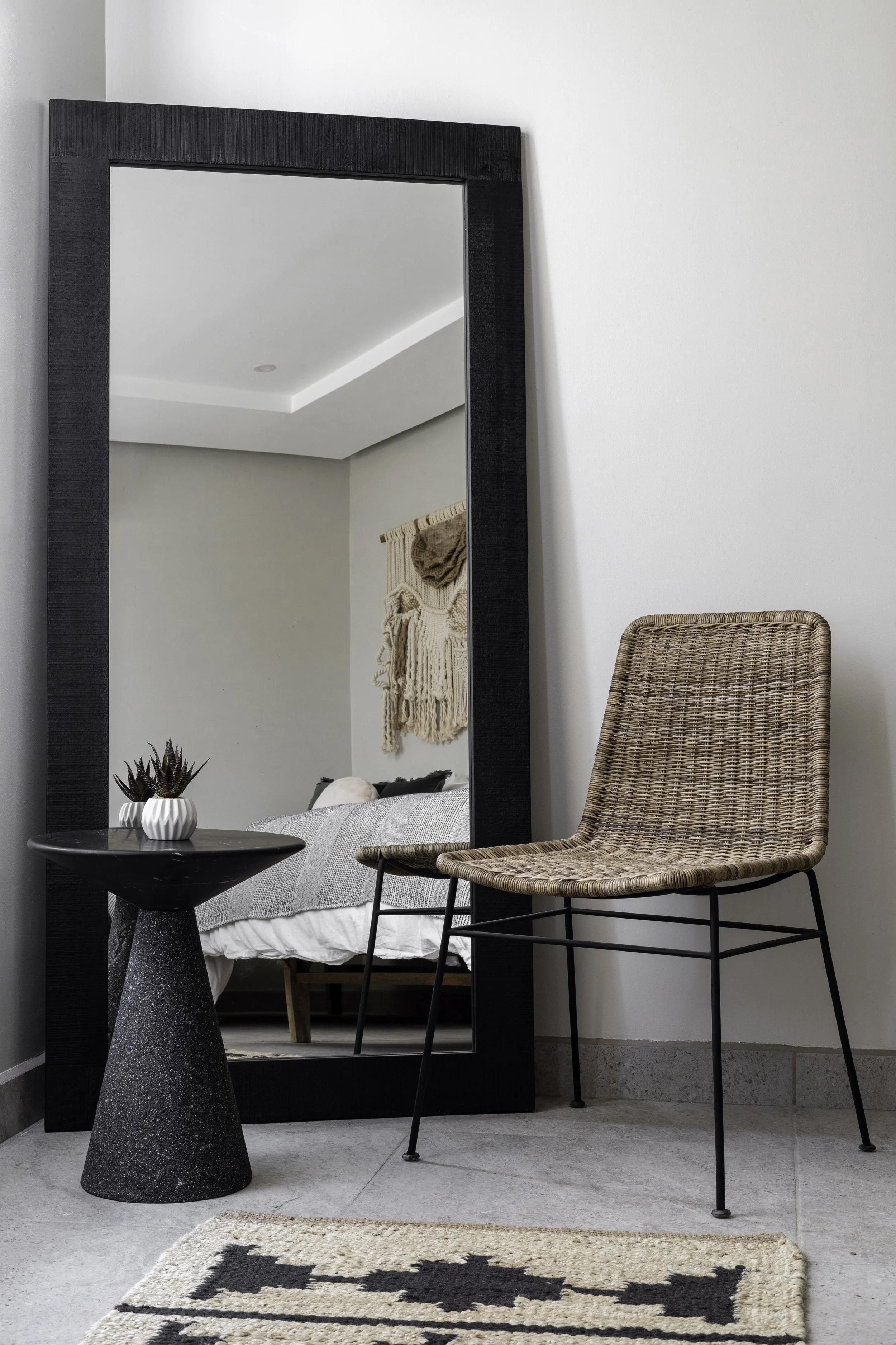 Stylish room with a rattan chair, black-framed mirror, small black side table, gray bedding, and decorative wall hanging.