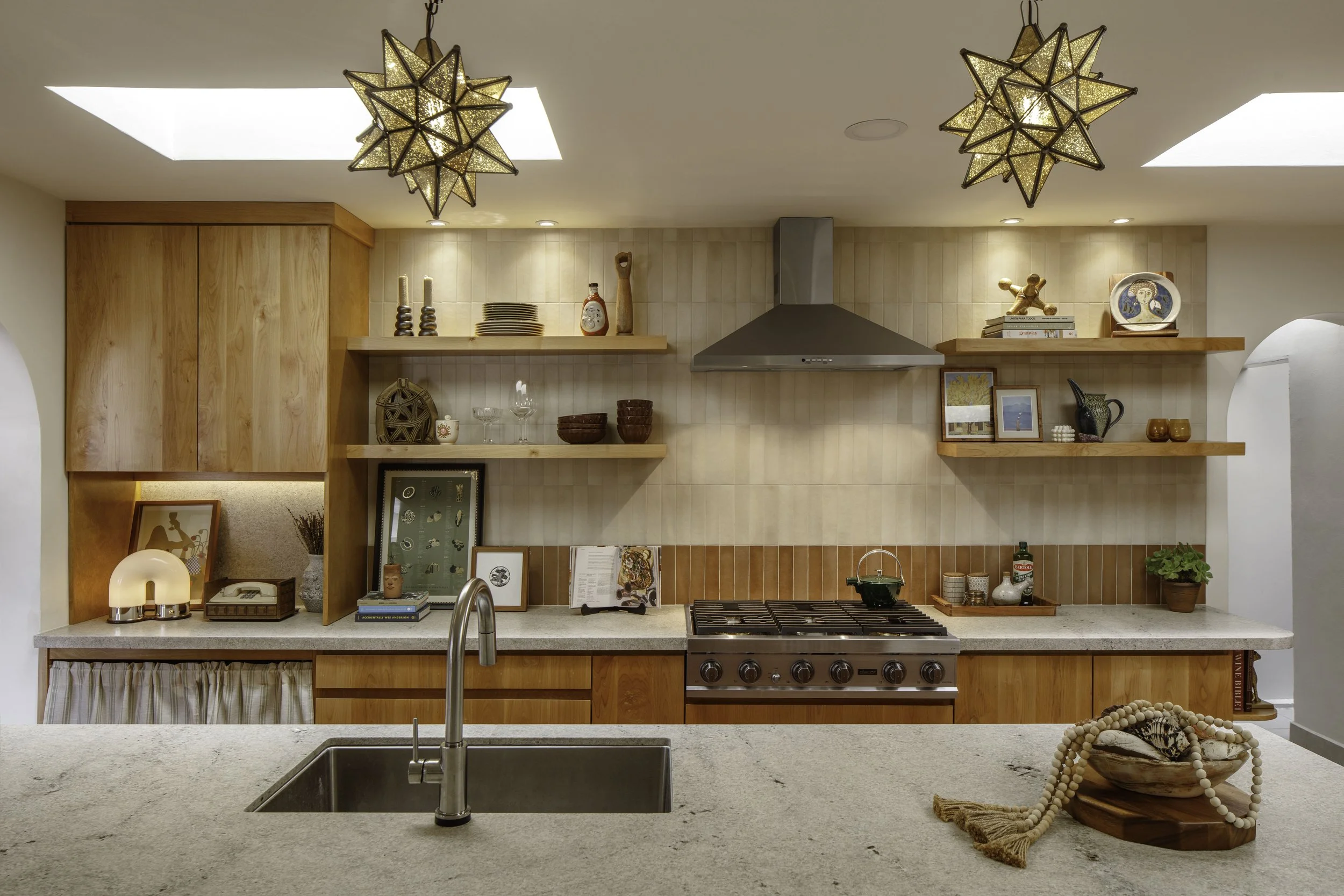 Modern kitchen with wooden cabinets, open shelves, and decorative items, featuring a stove with a metallic range hood, a beige countertop with a sink, and two geometric pendant lights hanging from the ceiling.