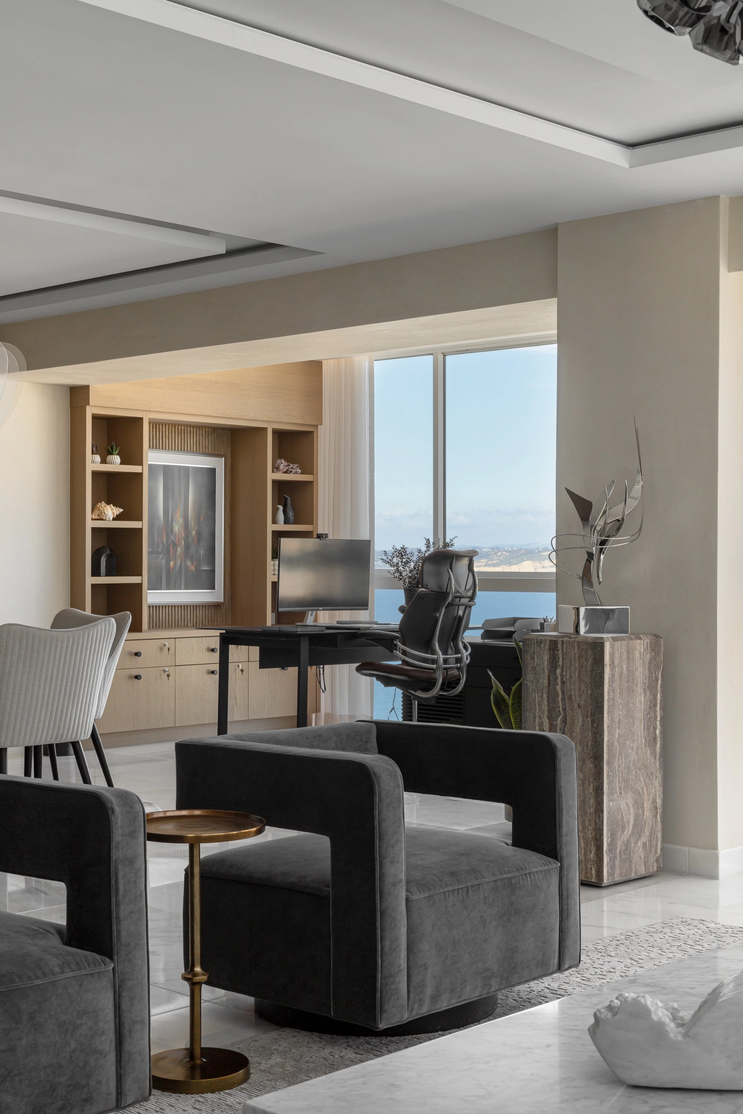 Modern living room with a home office setup by a large window. Includes a computer desk, ergonomic chair, shelves with decor items, and contemporary furniture. Ocean view visible through the window.