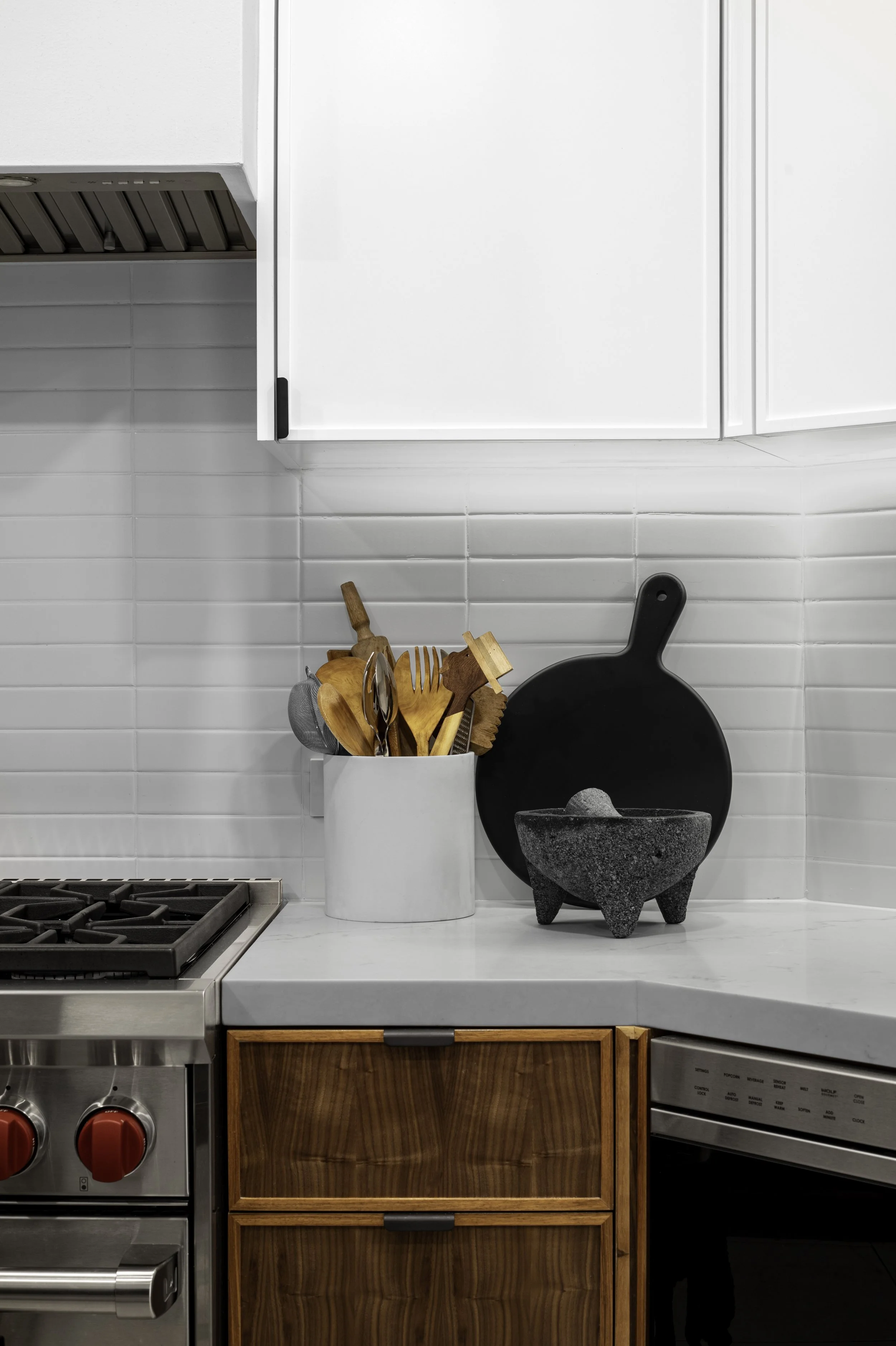 Modern kitchen corner with white cabinets, stainless steel stove, wooden drawers, and countertop accessories including a mortar and pestle, cutting board, and utensil holder.