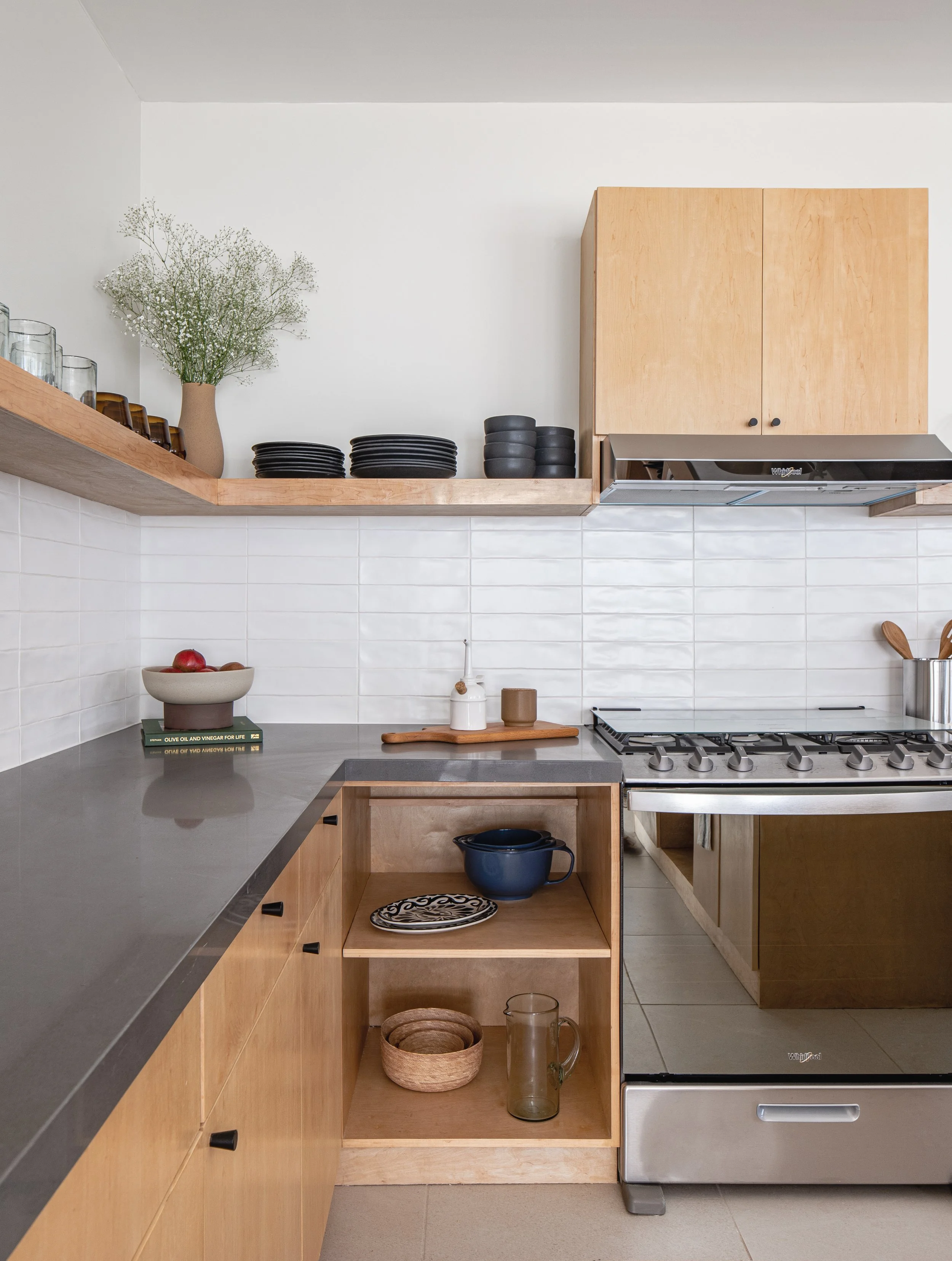 Modern kitchen with wooden cabinets, black countertop, gas stove, wall shelf with dishes, glassware, and plant in a vase.