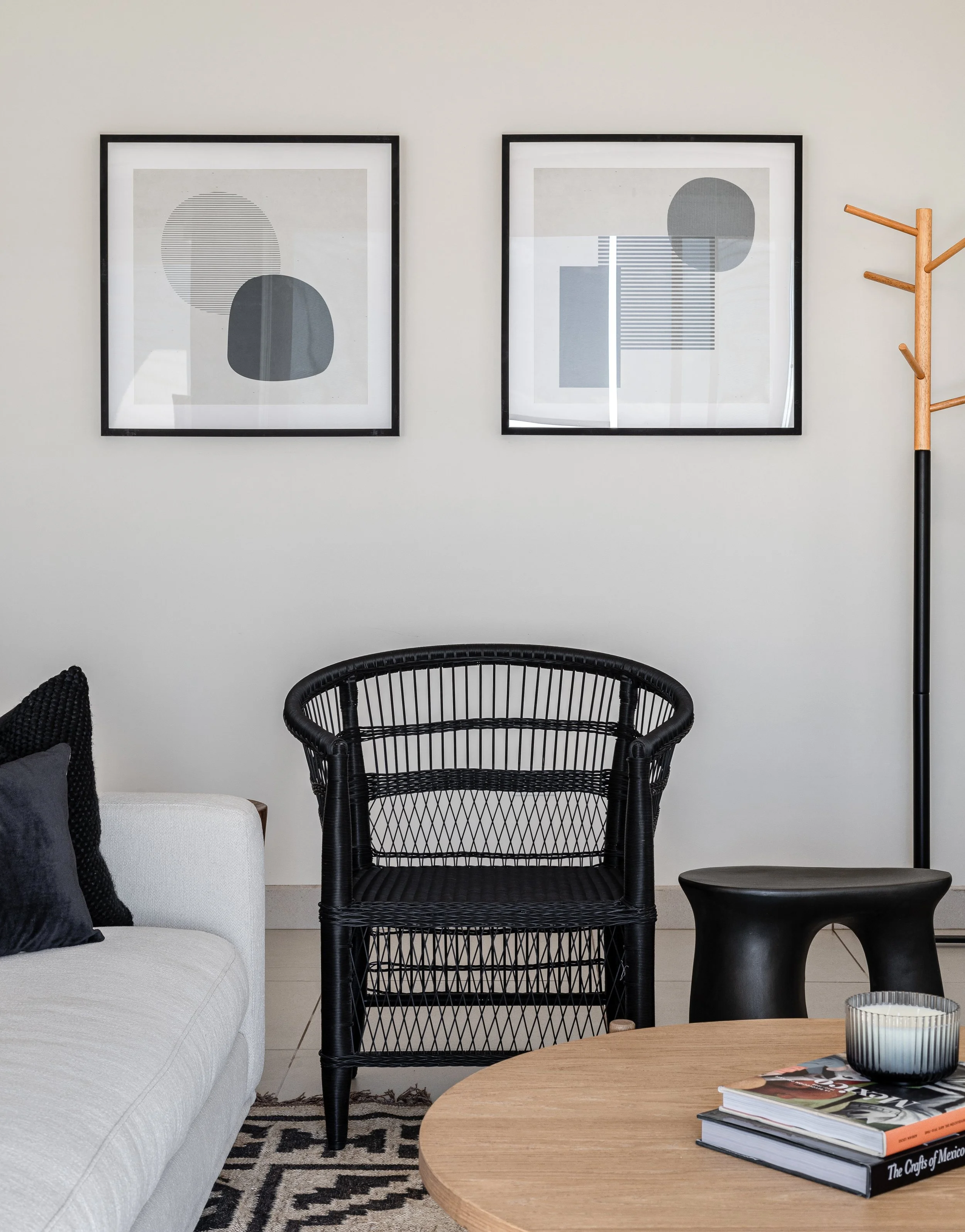 Modern living room interior with a black wicker chair, round wooden table, black stool, and light-colored sofa. Two abstract art pieces in black frames hang on the wall above. A black and white patterned rug covers the floor. A wooden coat rack is vi