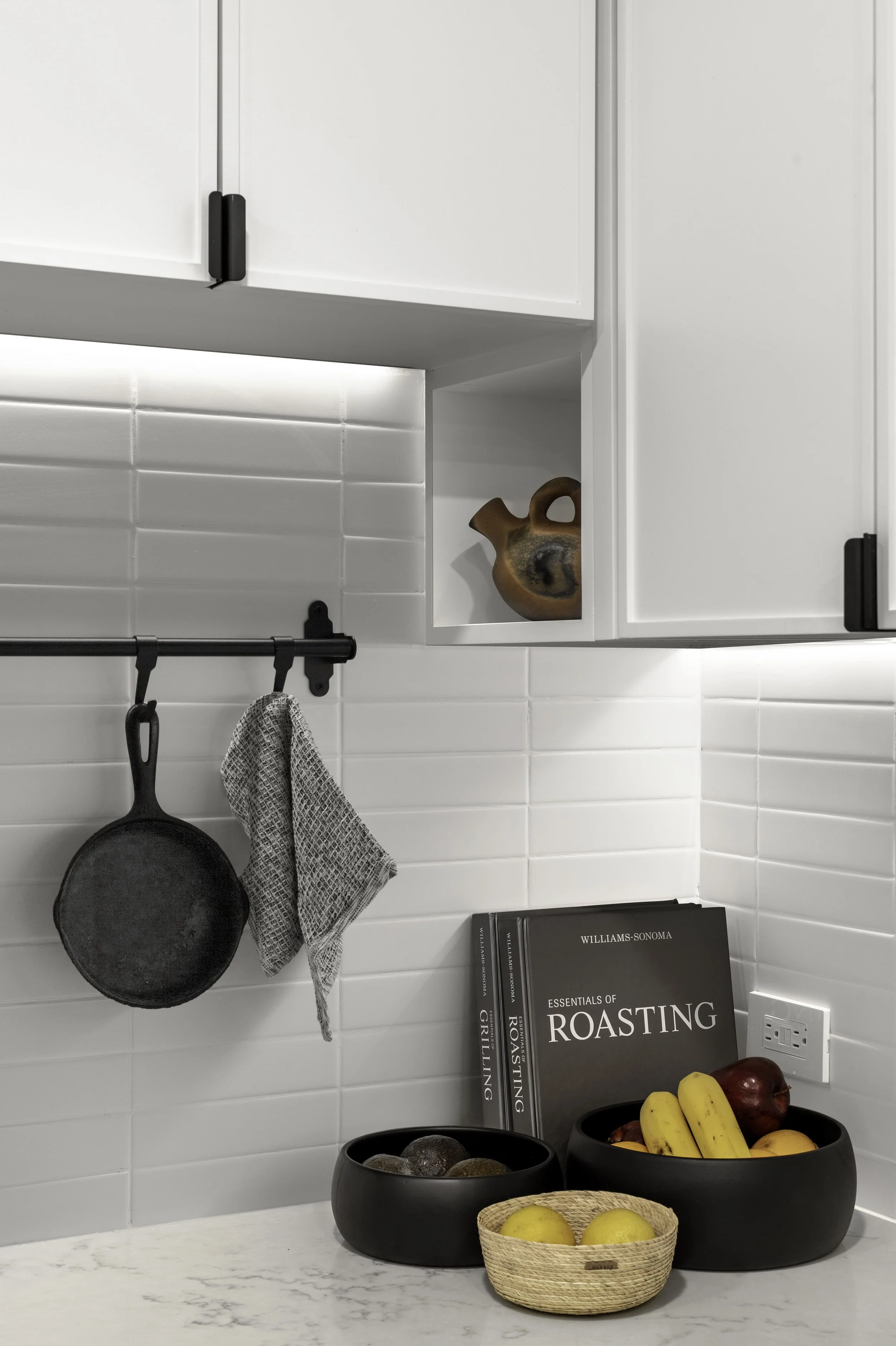 Modern kitchen corner with white tile backsplash, white cabinets, a black cast iron skillet, a gray hand towel, decorative pottery, cookbooks including "Essentials of Roasting," and black bowls with fruits, on a marble countertop.