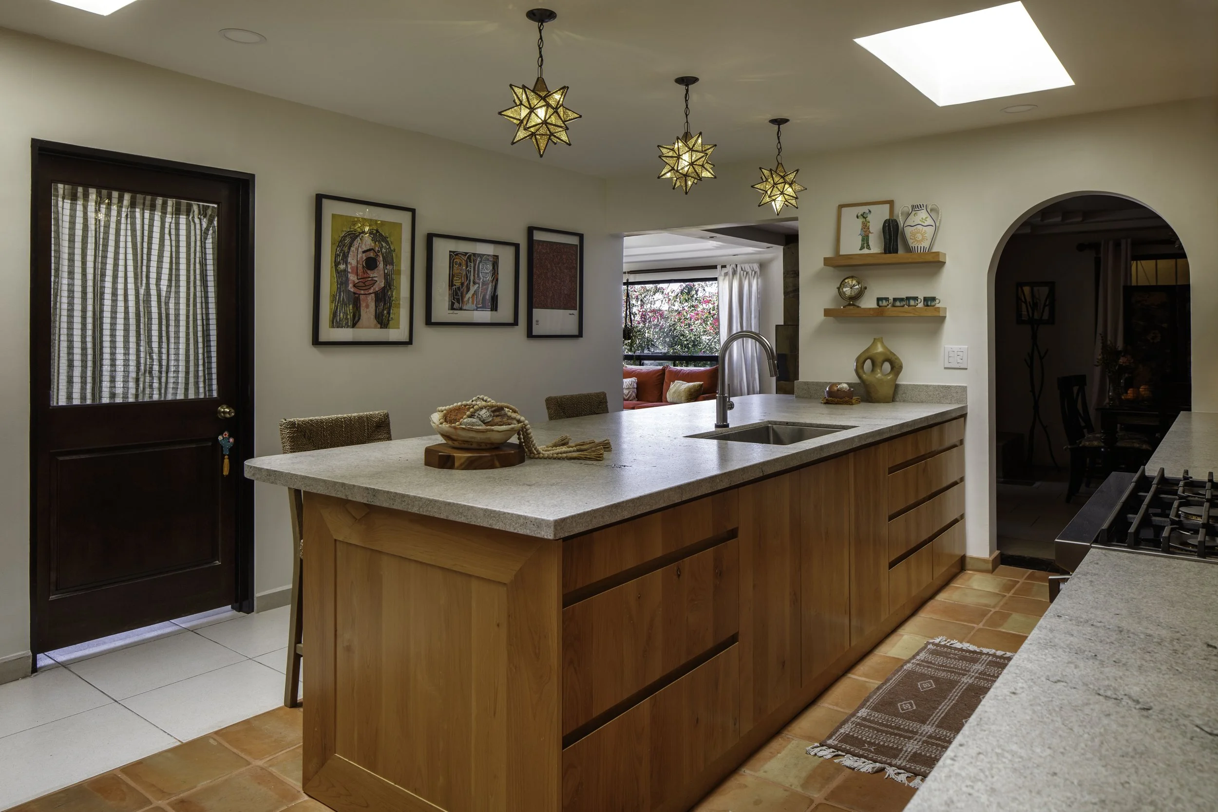 A modern kitchen with a central island, wooden cabinets, and a gray countertop. There are three star-shaped pendant lights hanging from the ceiling, a skylight, and artwork on the walls. The space opens into a dining area with plants outside the wind