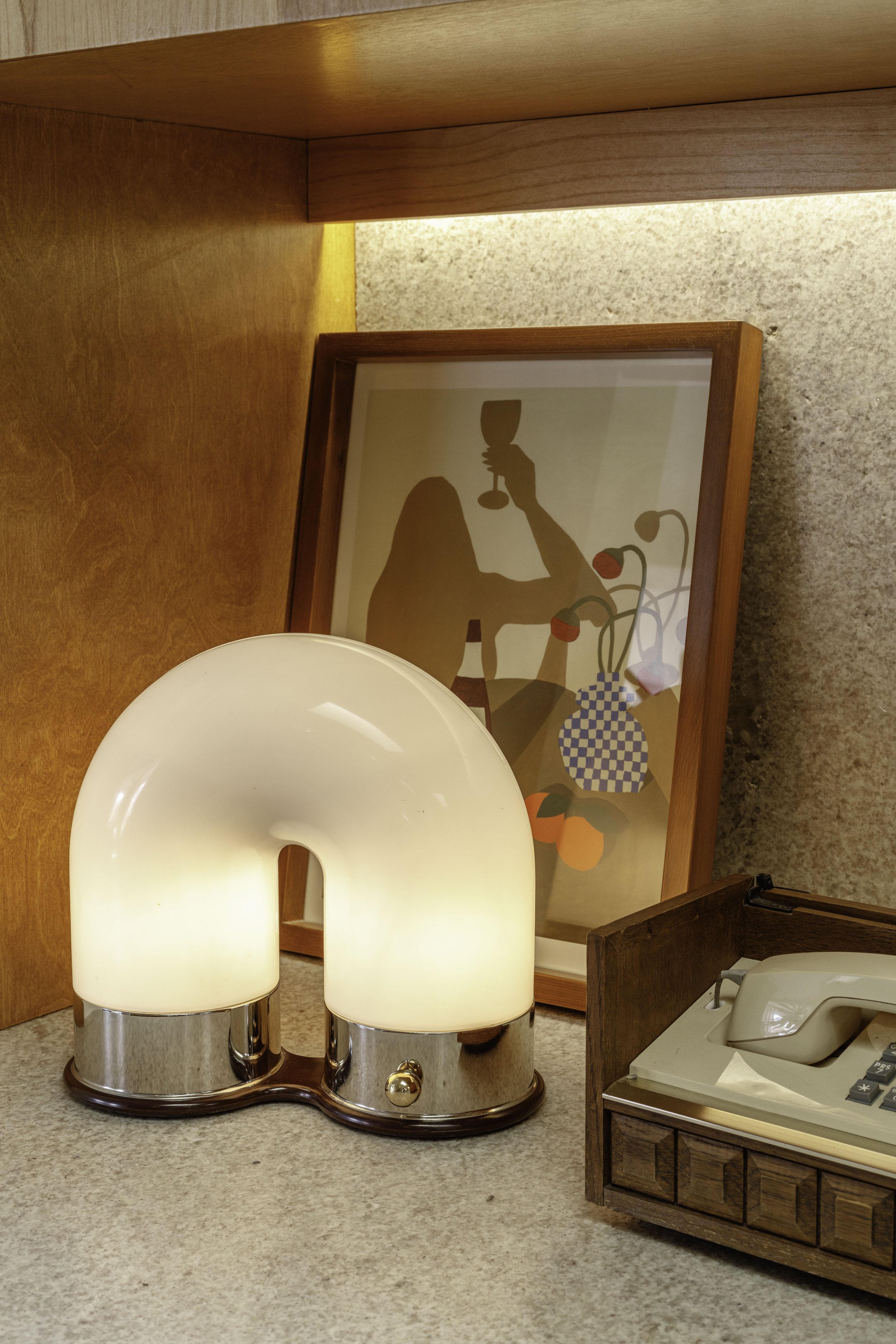 A vintage table lamp with a curved white shade and a chrome base, placed on a beige countertop next to a framed abstract art print and a wooden box holding a vintage white landline telephone.