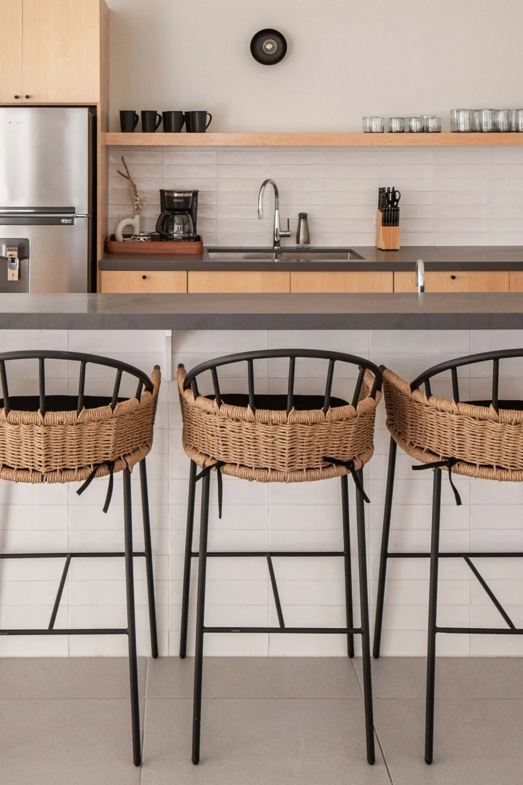 Modern kitchen interior with stainless steel appliances, three wicker bar stools, black and wooden elements, and gray countertops.