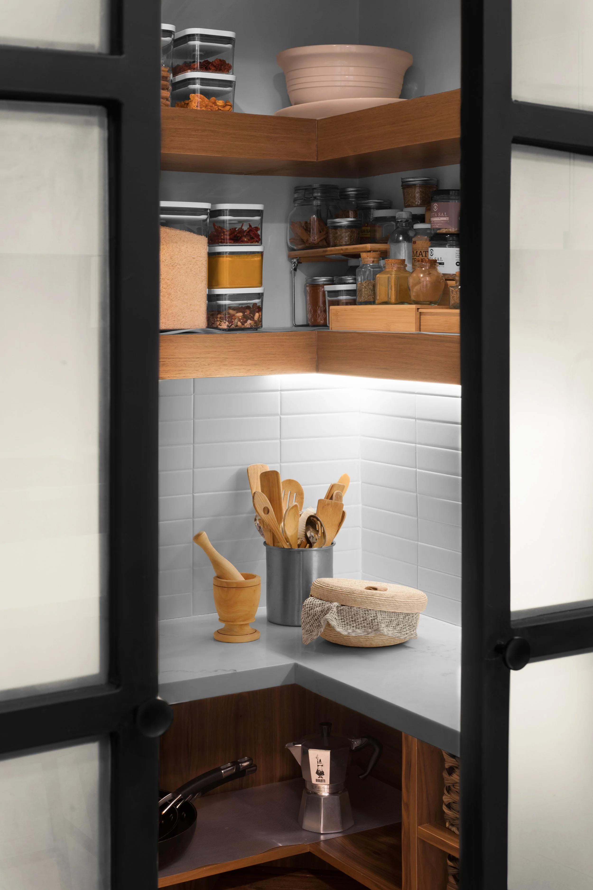 Modern pantry with wooden shelves and various jars containing spices, grains, and dried goods, along with kitchen utensils in a metal holder, a mortar and pestle, and a wicker basket on the countertop.