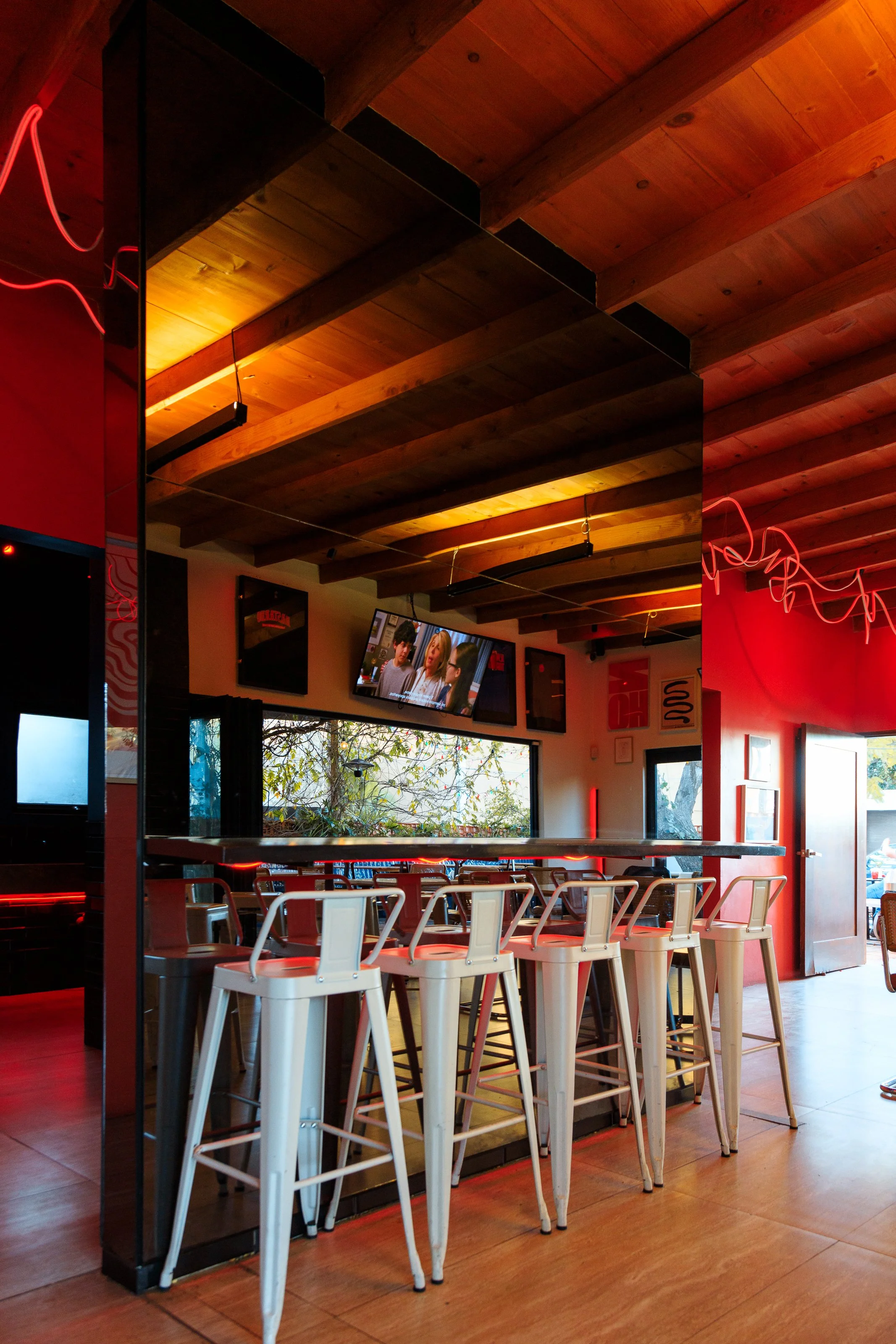 Interior of a modern bar with high stools, a wooden ceiling, and a wall-mounted TV displaying a show, featuring red and orange ambient lighting.