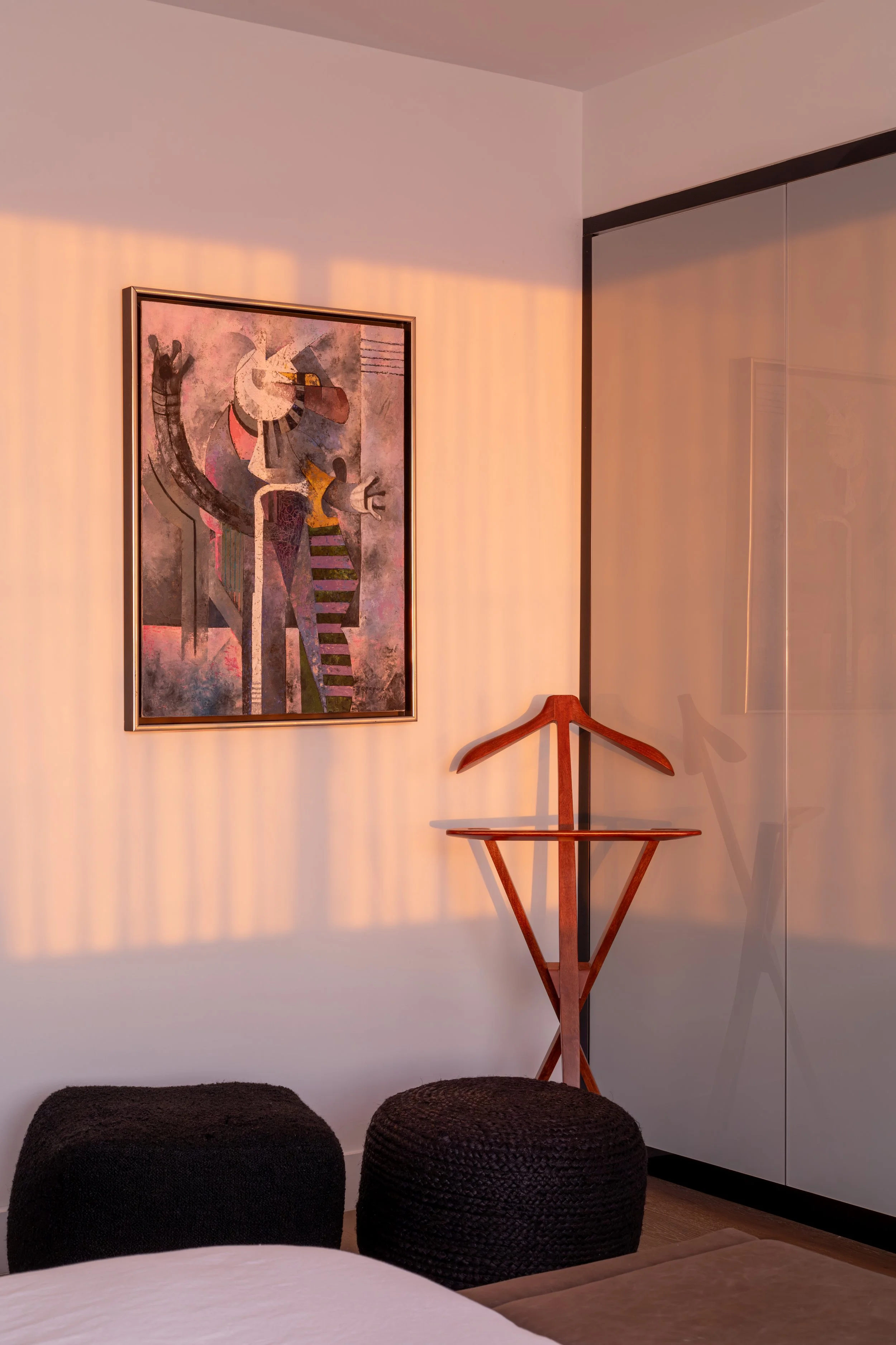 Two black woven poufs, a wooden valet stand, and an abstract painting on a wall in a room with warm sunlight.
