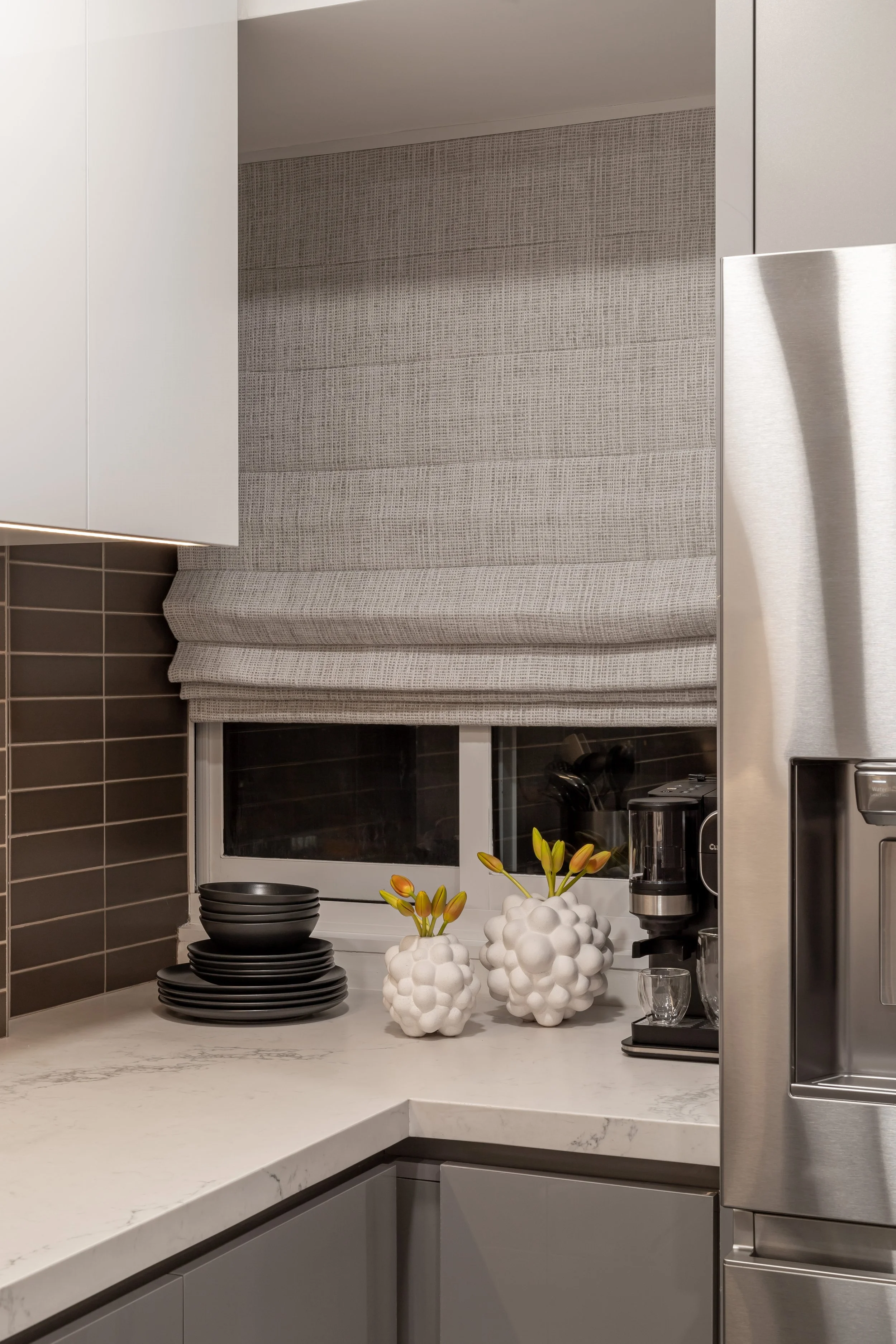 Modern kitchen corner with white countertop, gray Roman shade blind, stack of black plates, white geometric vases with yellow flowers, espresso machine, and stainless steel appliances.