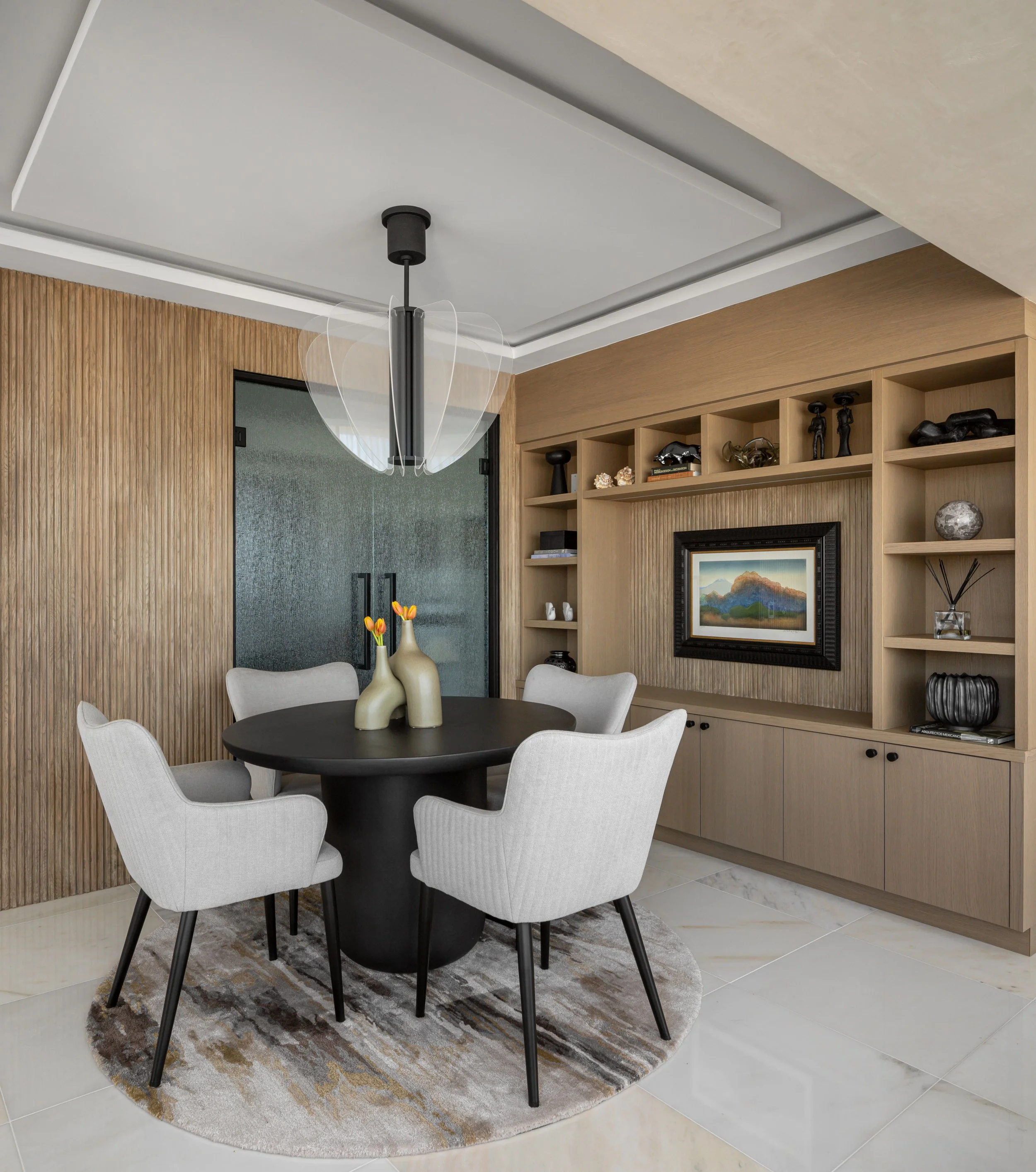 Modern dining room with round black table, four gray chairs, a circular rug, and a light fixture. Shelving units with decor items and a framed painting adorn the wall.