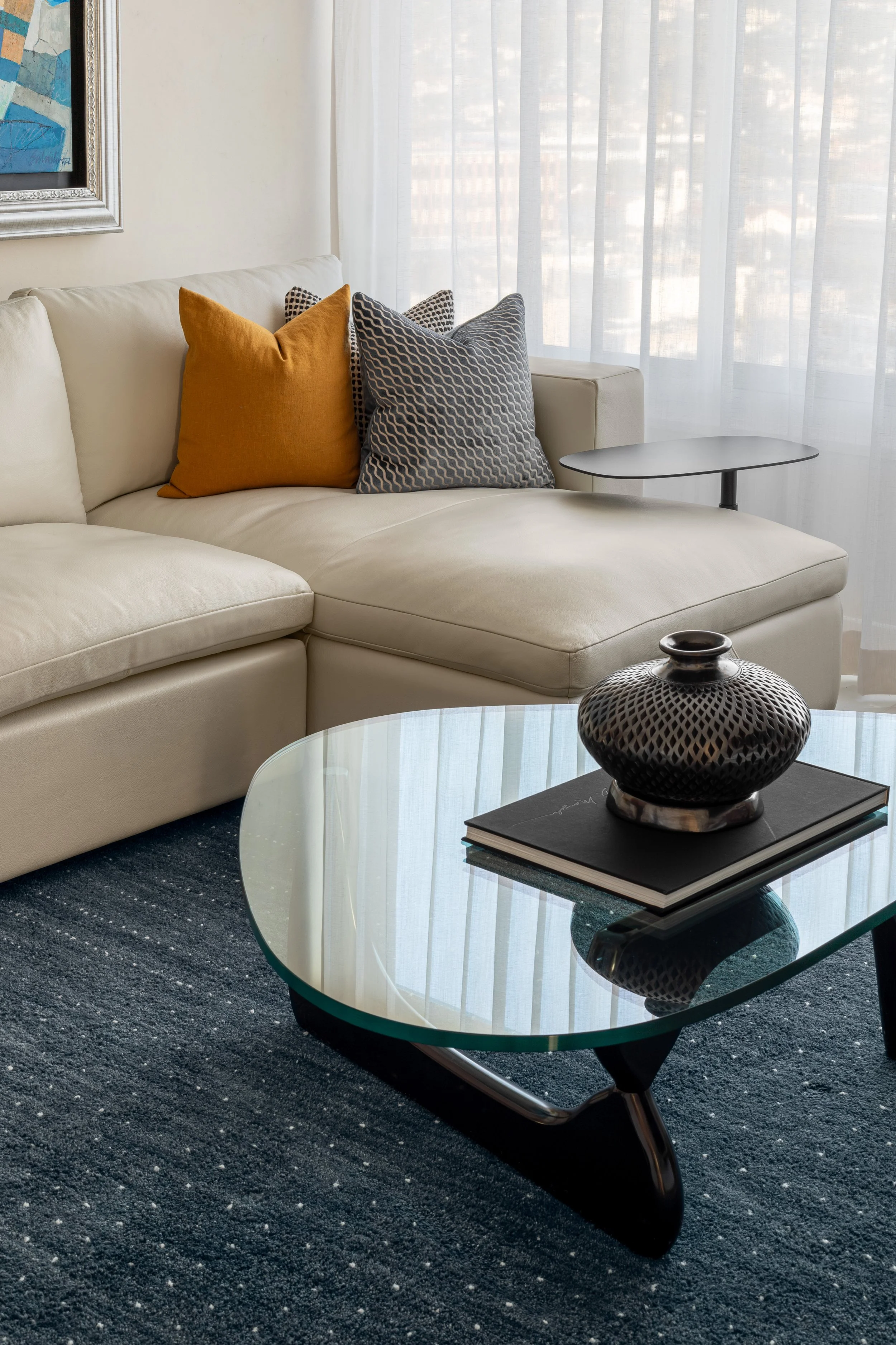 Living room with a white sofa, yellow and patterned pillows, glass top coffee table with a black vase and a notebook, dark textured rug, side table, and sheer curtains.