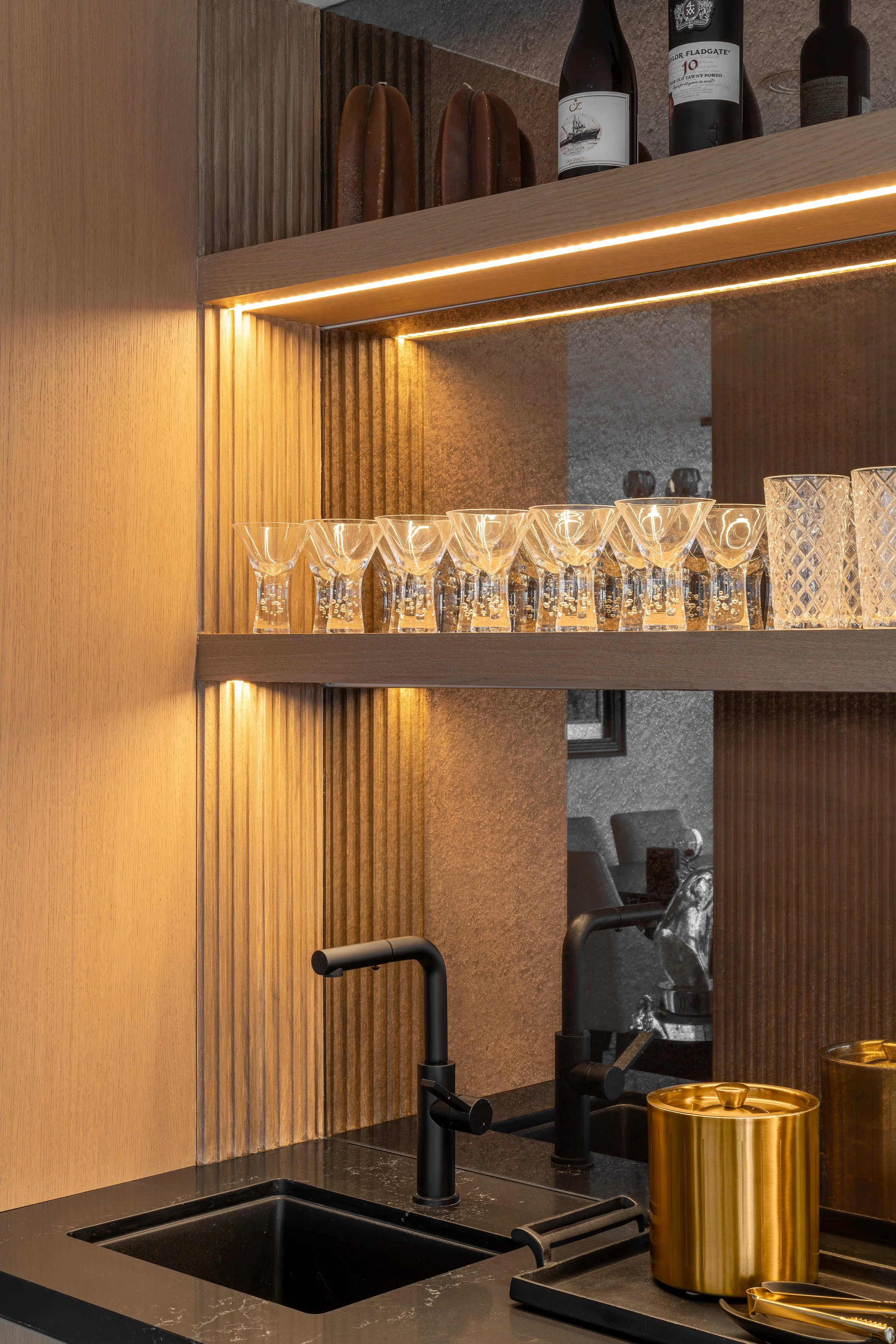 Modern kitchen counter with black sink, gold accents, glassware and wine bottles on wooden shelves, illuminated by warm lighting.