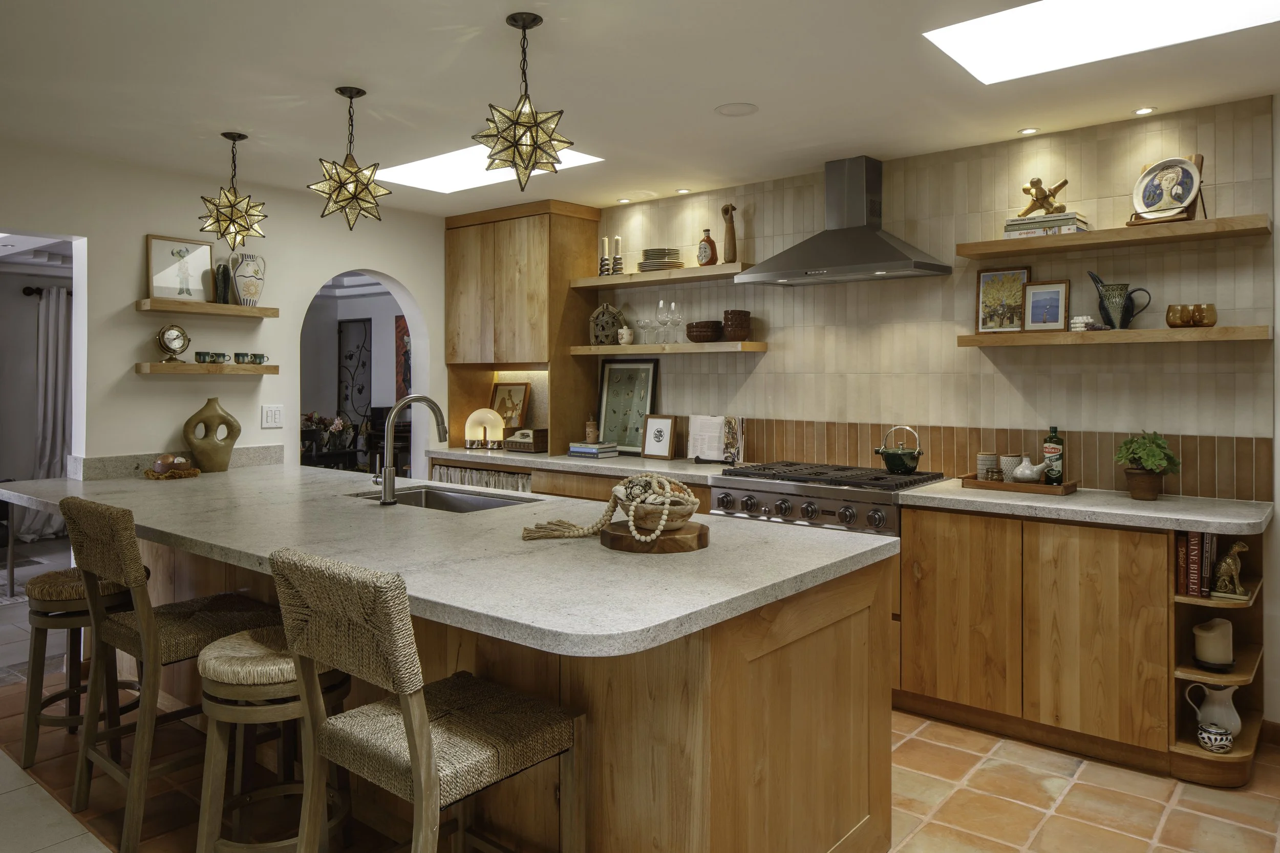 Modern kitchen with wooden cabinets, open shelves decorated with art and dishware, a gray countertop island with wicker chairs, star-shaped pendant lights, and a tiled backsplash.