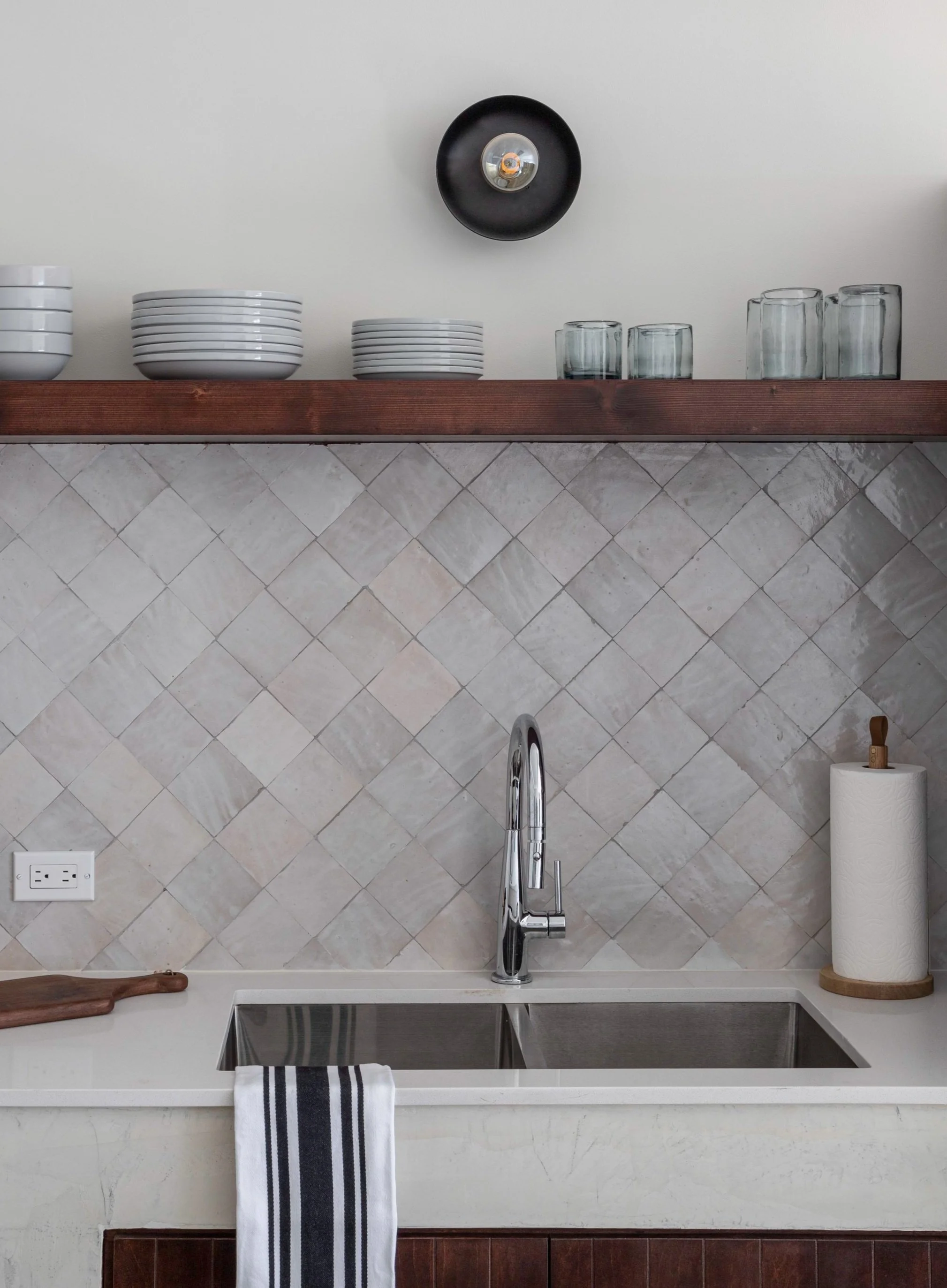 Modern kitchen sink area with a stainless steel faucet, white countertops, and a light gray tiled backsplash. Above is a wooden shelf holding stacked plates and glasses. A paper towel holder and striped dish towel are on the counter. A round black li