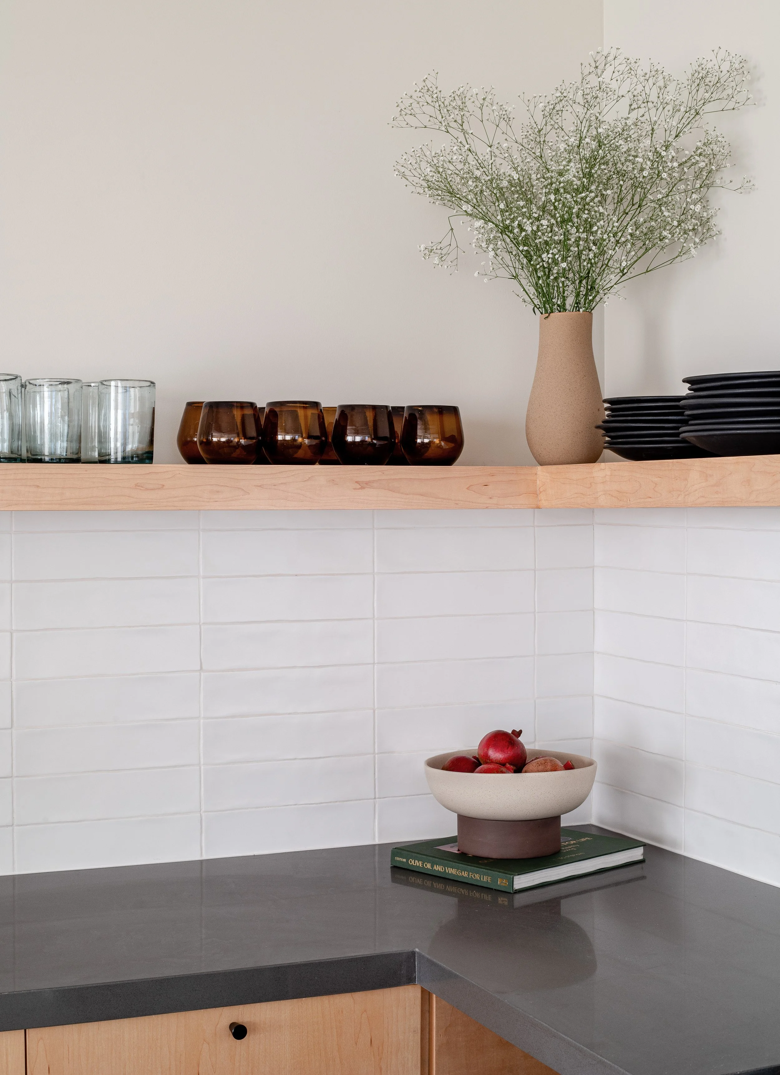 Modern kitchen with glassware and potted flowers on shelves, white tiled walls, and a counter holding a bowl of pomegranates on a stack of books.