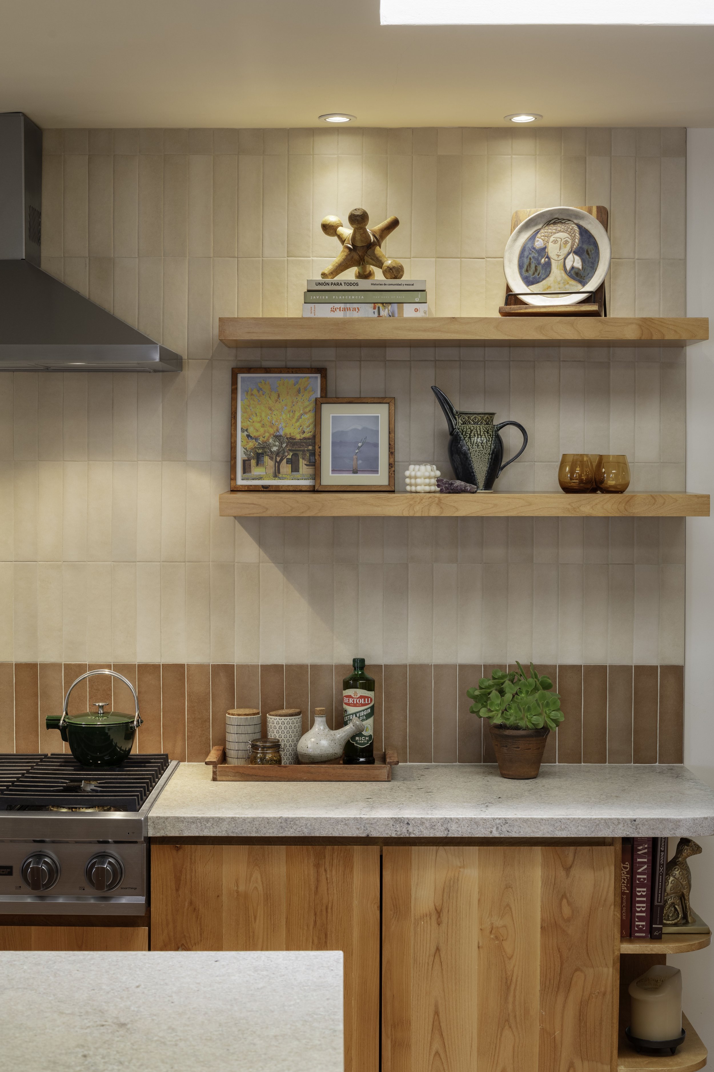 A kitchen shelf with decorative items, framed paintings, ceramic and glassware, and a potted plant above a countertop with jars, a bottle of olive oil, and a green plant in a terracotta pot.