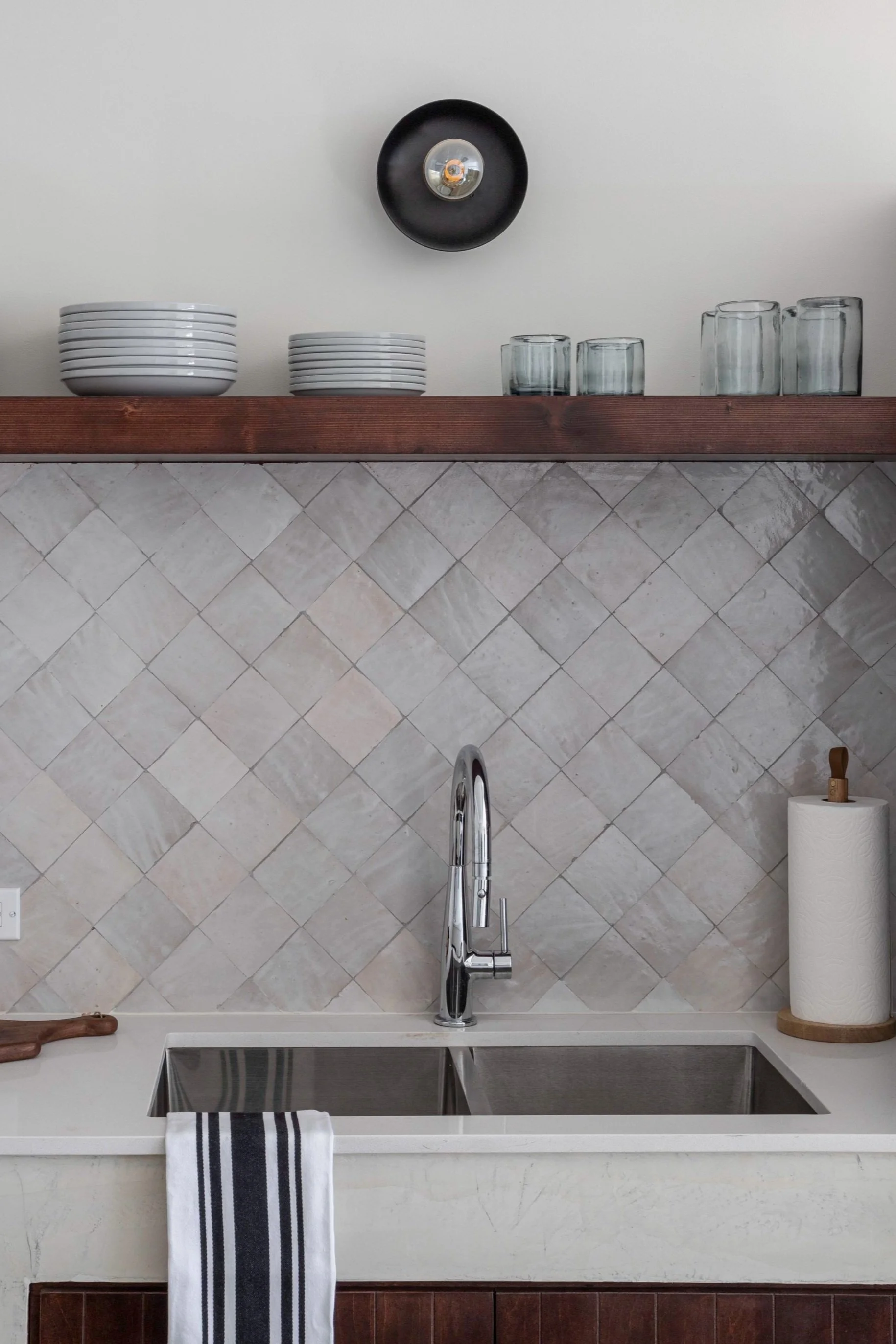Kitchen with a stainless steel double sink, a chrome faucet, a paper towel roll on a wooden holder, a dish towel hanging, a backsplash of beige tiles, a wooden shelf with bowls and glasses, and a black wall sconce