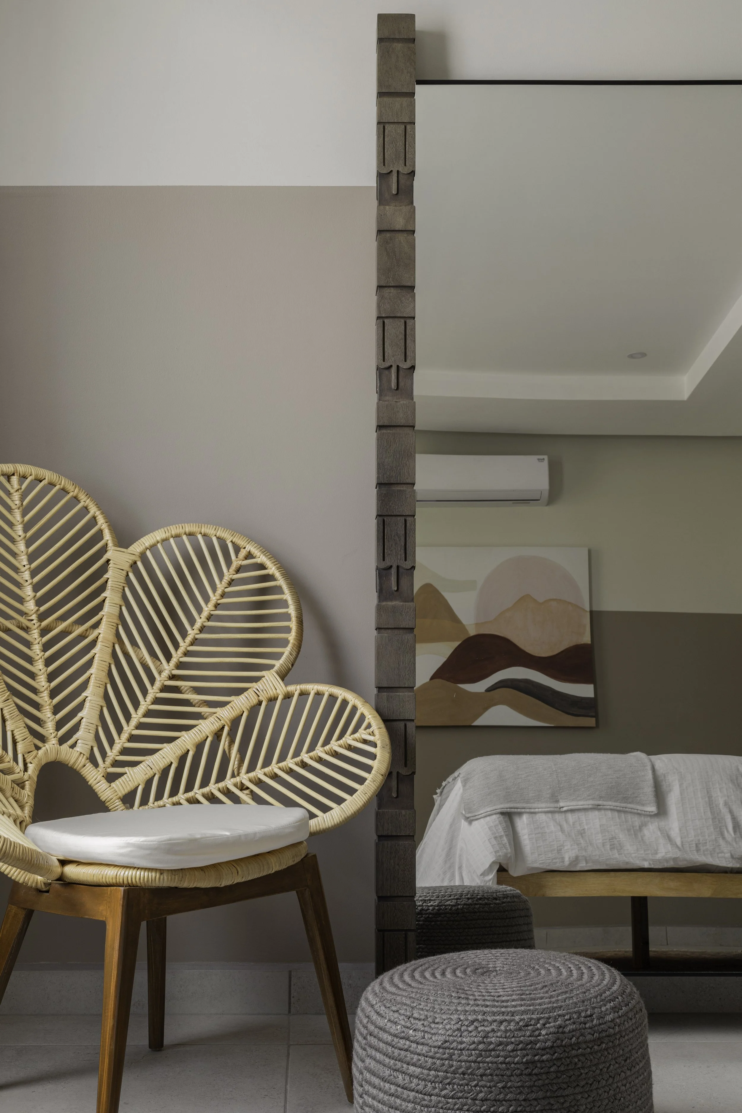 Interior view of a room with a rattan chair, a textured gray ottoman, partial view of a bed with white bedding, a painting of mountains, and a decorative room divider with a brown textured design.