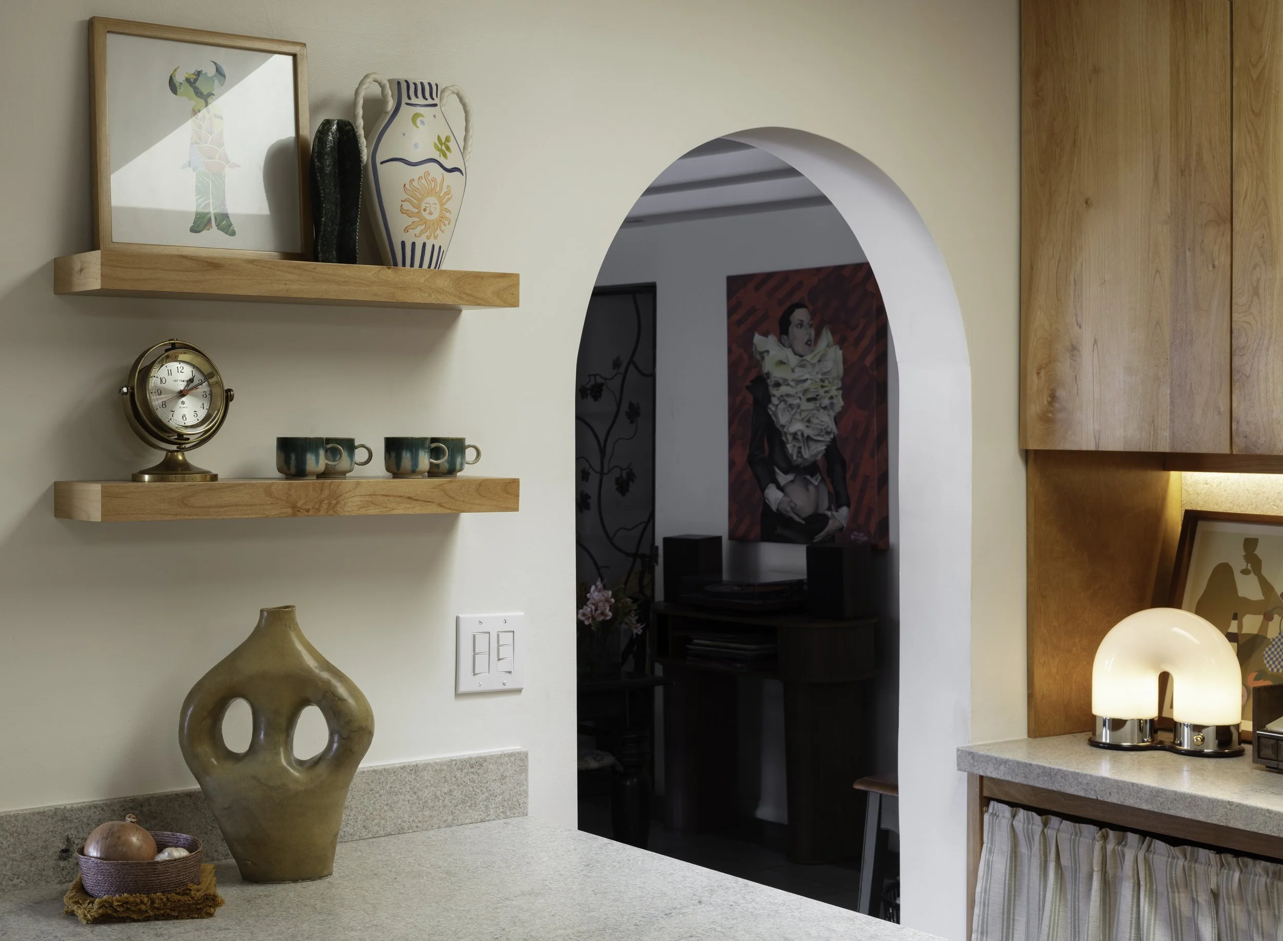 Part of a kitchen with two wooden shelves holding decorative items, a vintage clock, and three coffee mugs. A large ceramic vase is on the countertop, and decorative objects under a lamp on the right. An arched doorway reveals a room with artwork and