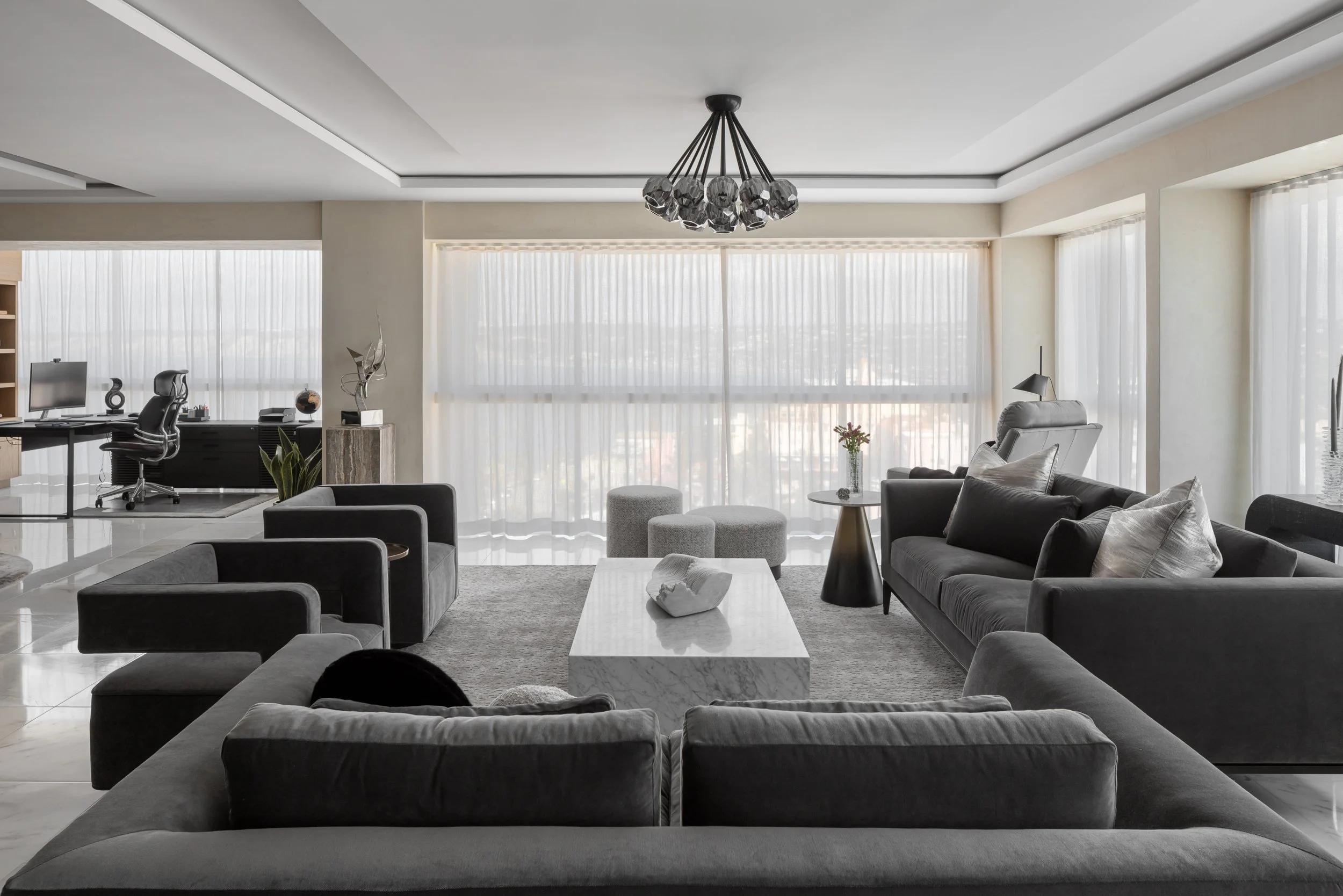 Modern living room with gray sofas, coffee table, and a desk with computer, next to large windows with sheer curtains.
