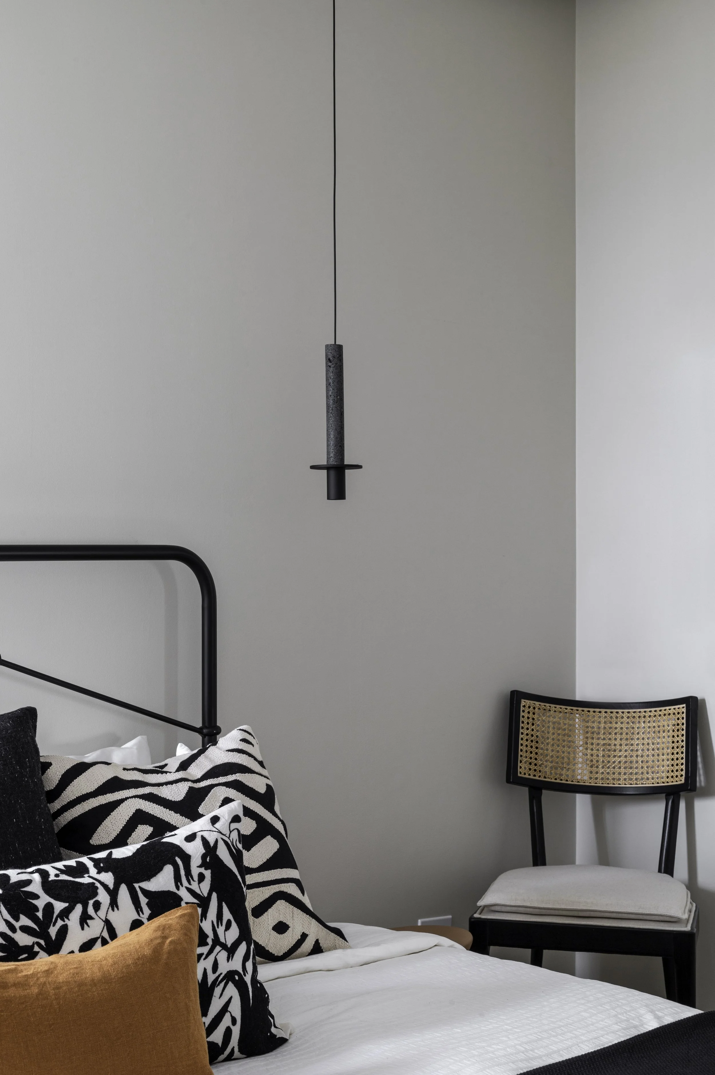 Minimalist bedroom with a bed featuring geometric and floral pillows, black metal headboard, and a cane-back chair. A pendant light hangs above.