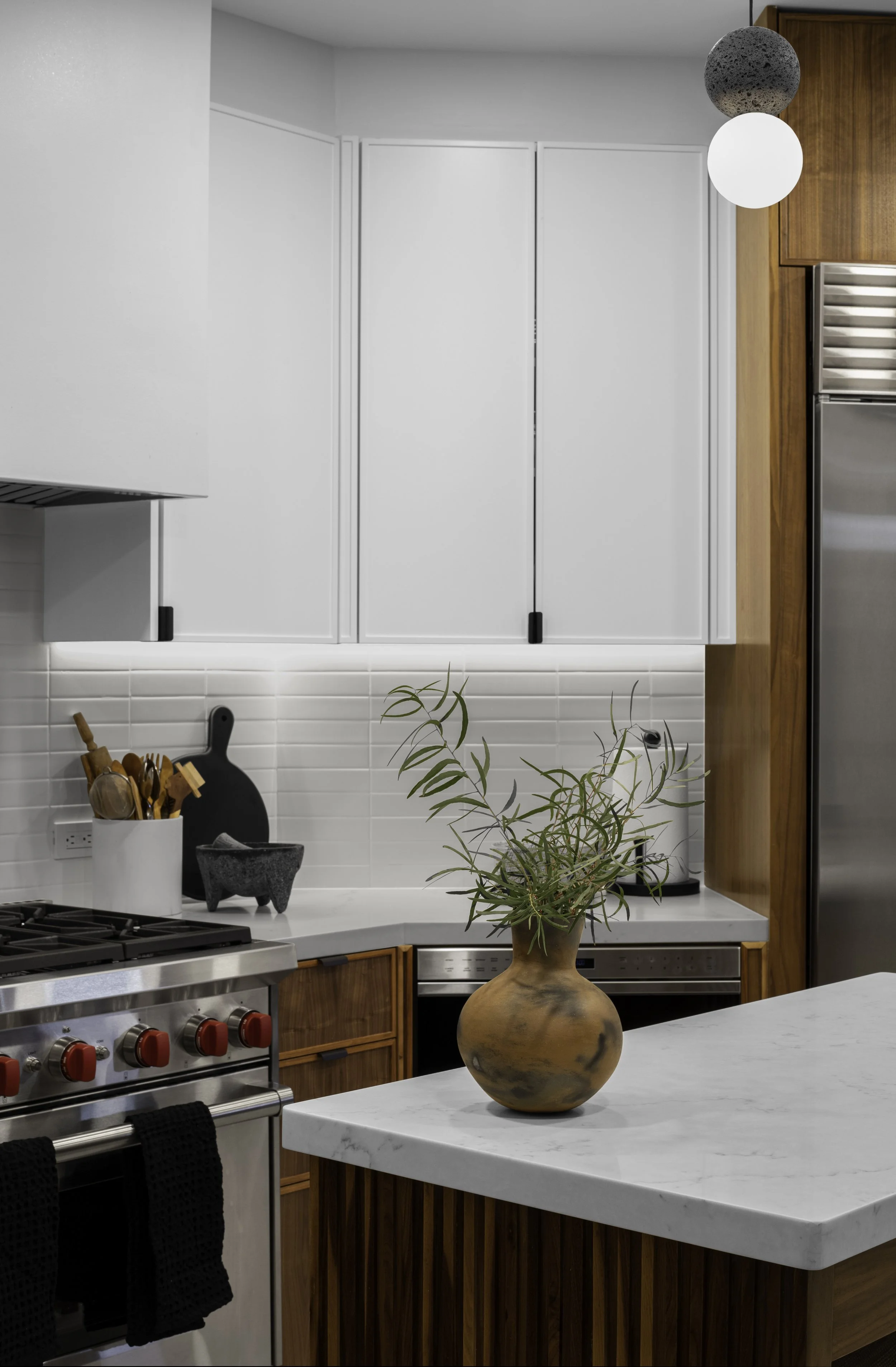 Modern kitchen with stainless steel stove, white cabinetry, potted plant on marble countertop, wooden accents, and decorative utensils in a holder.