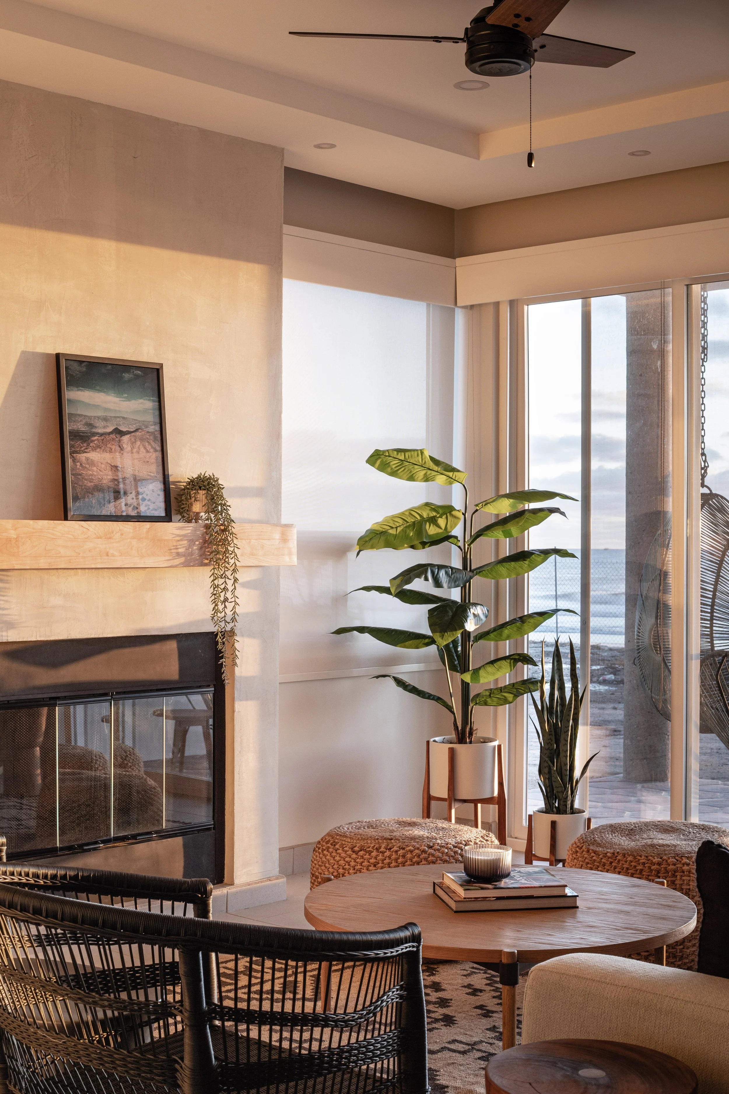Cozy living room with modern decor, featuring a fireplace with a framed photo on the mantel, houseplants, wicker chairs, poufs, and a wooden coffee table with books.