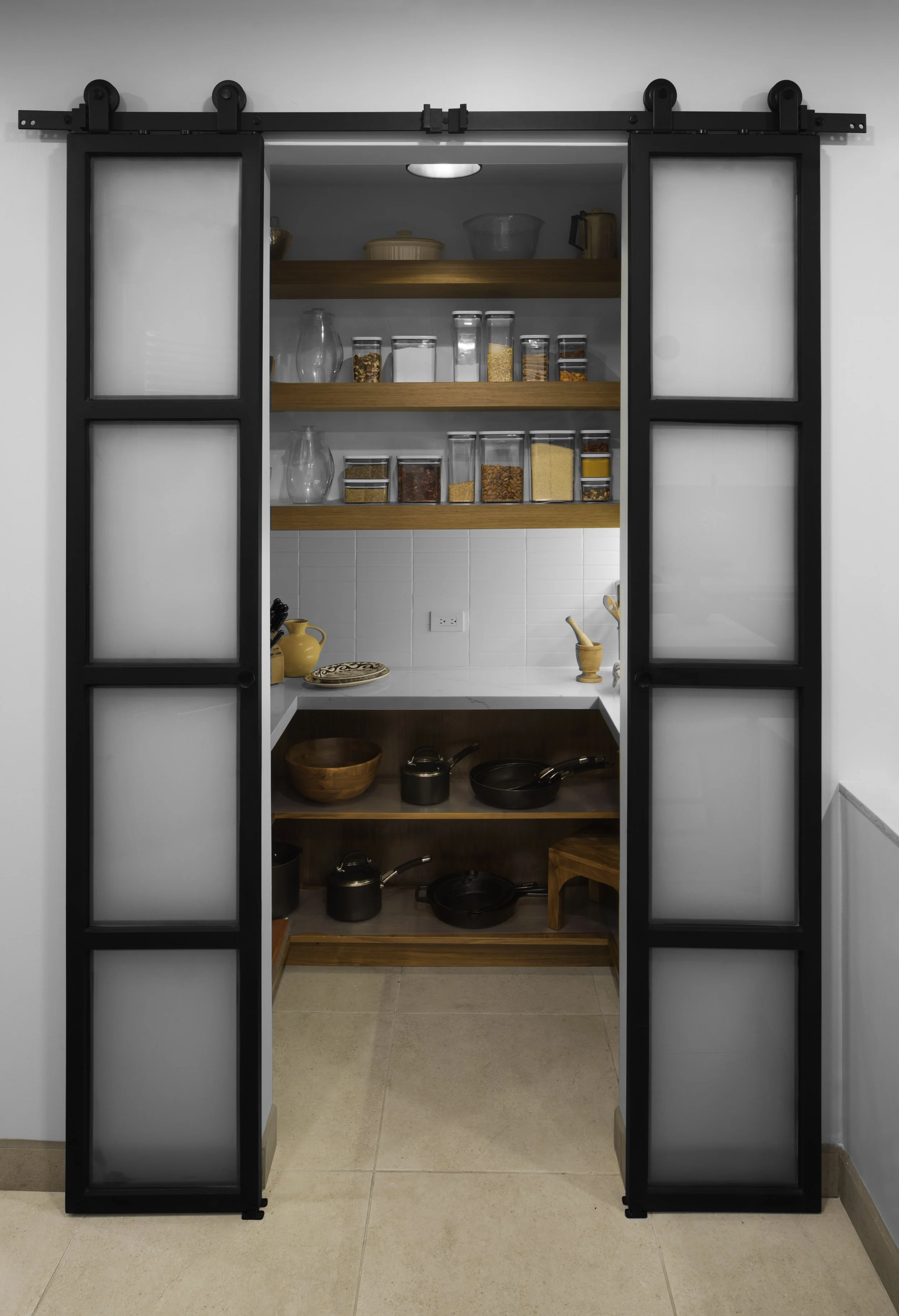 Sliding pantry door with frosted glass panels, wooden shelves with glass jars of food, pots and pans on lower shelves, white tiled backsplash.