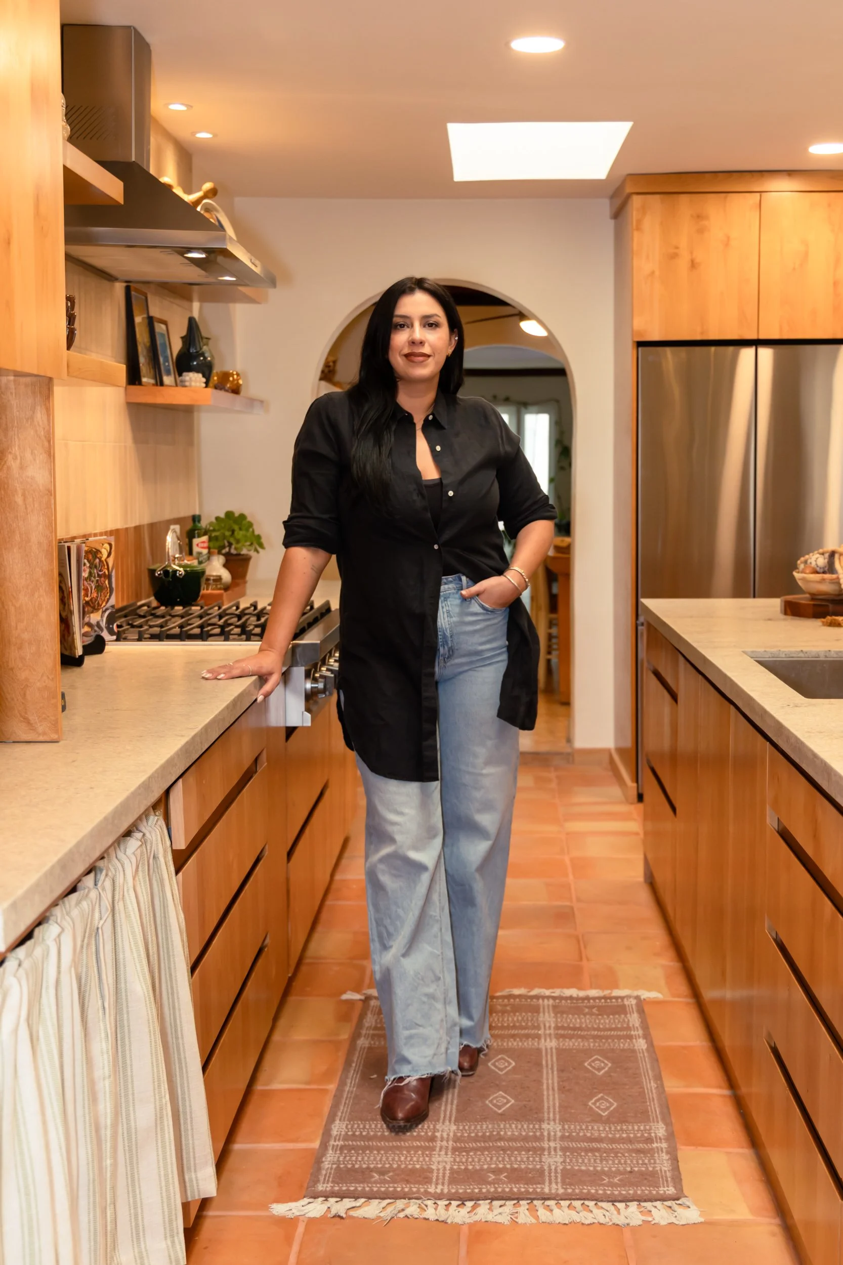 A woman with long black hair wearing a black long shirt, light blue jeans, and brown boots standing in a kitchen with wooden cabinets and a tile floor.