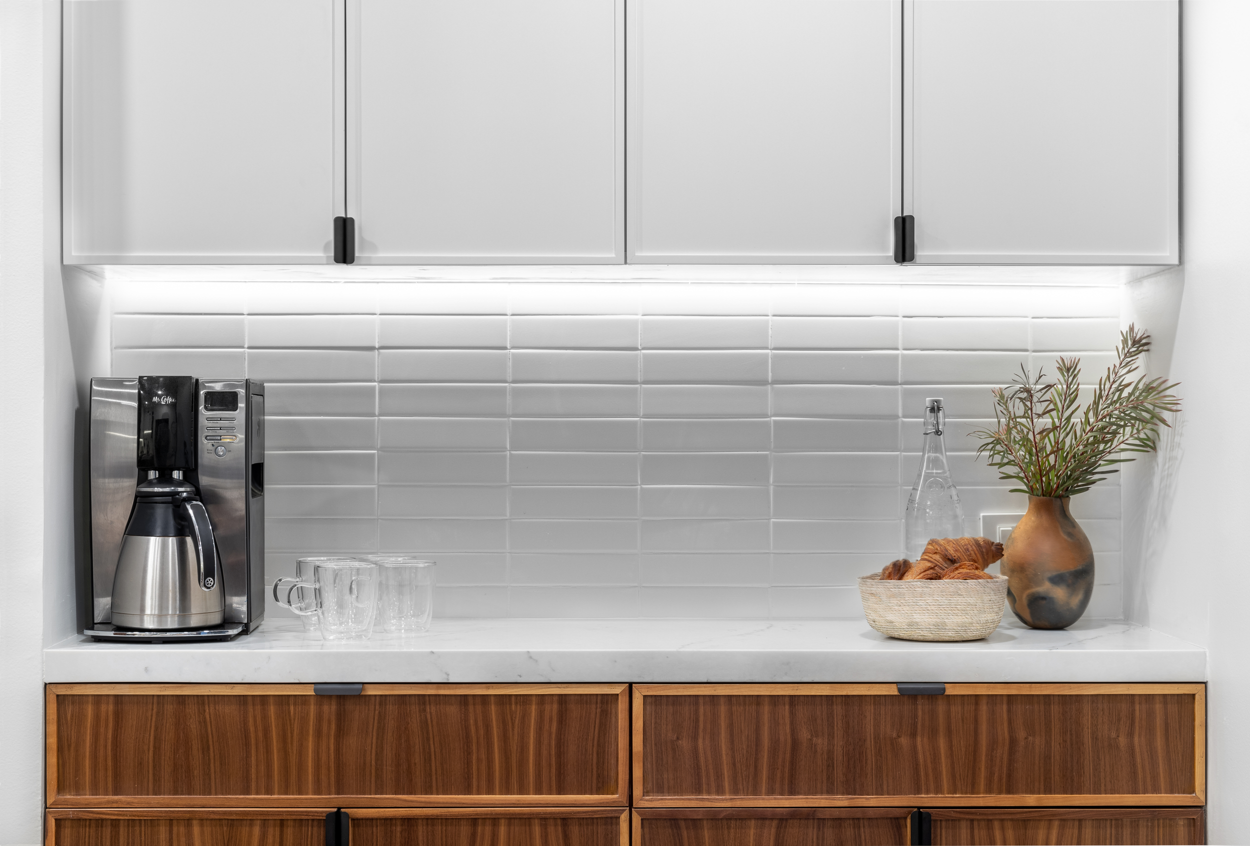 Modern kitchen countertop with a coffee maker, glass mugs, a basket of croissants, a water bottle, and a vase with greenery. White cabinets and tiled backsplash above wooden drawers.
