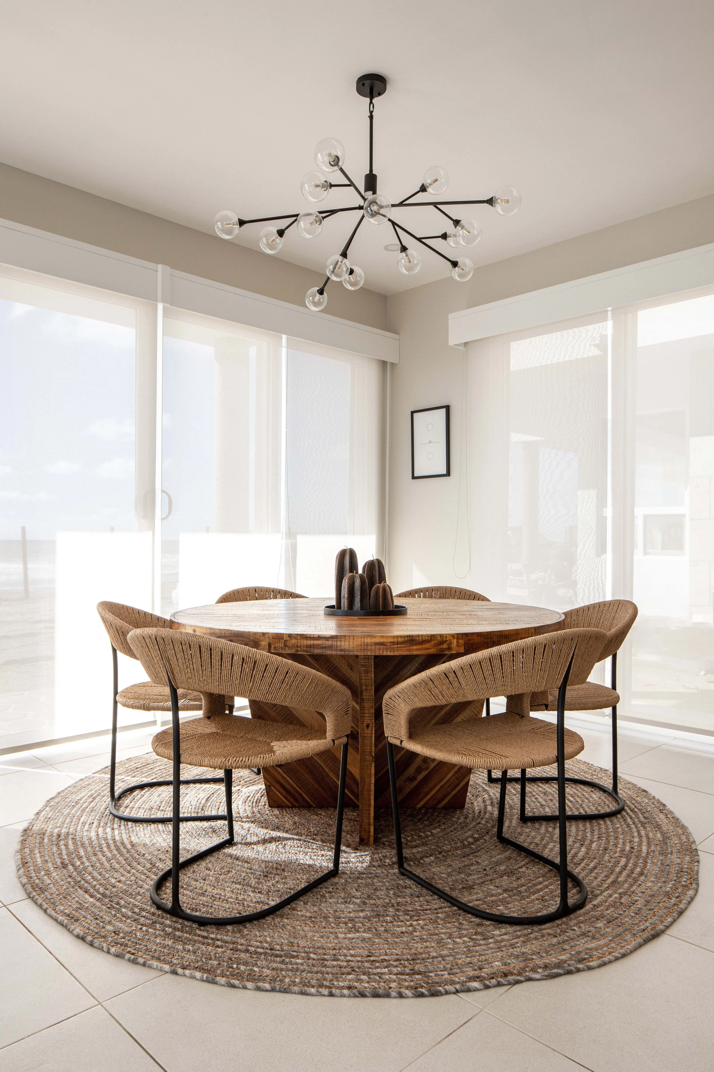 Modern dining area with a round wooden table, surrounded by wicker chairs on a woven rug. A contemporary chandelier hangs above, and large windows offer natural light.