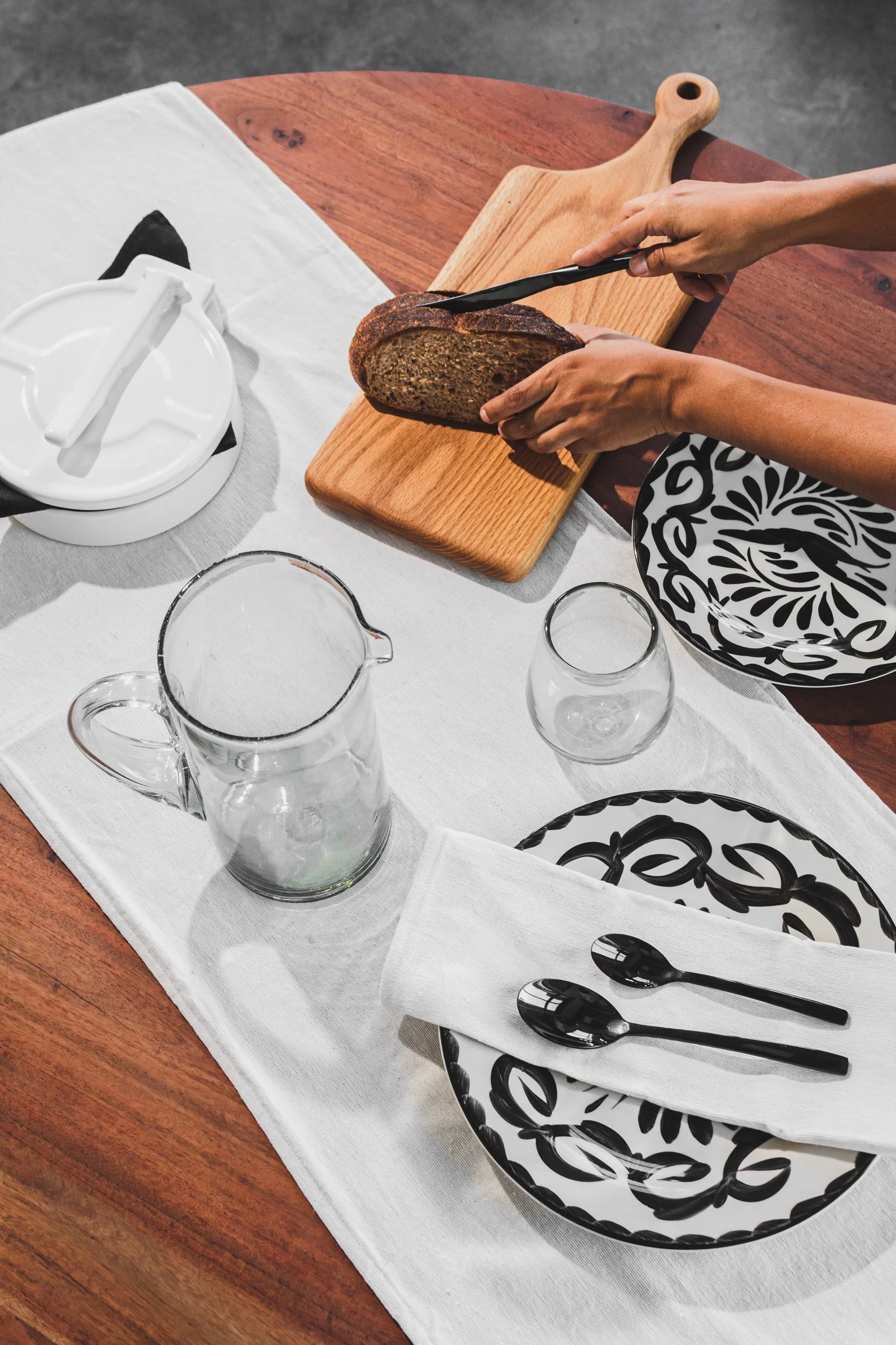 Person slicing a loaf of bread on a wooden cutting board at a dining table with black and white patterned plates, a white linen table runner, and glassware.