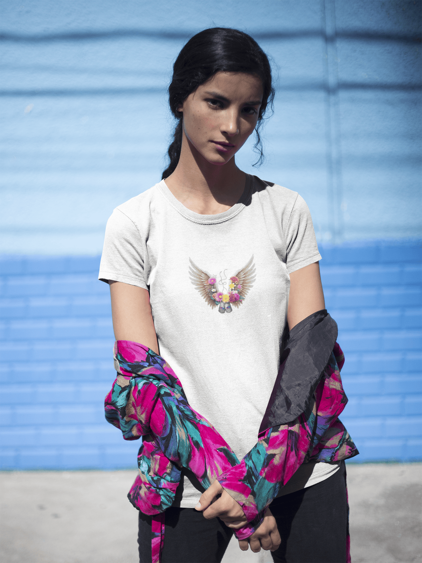 A woman with short dark hair and multiple tattoos wearing a black t-shirt with a colorful angel wings design and loose blue jeans.