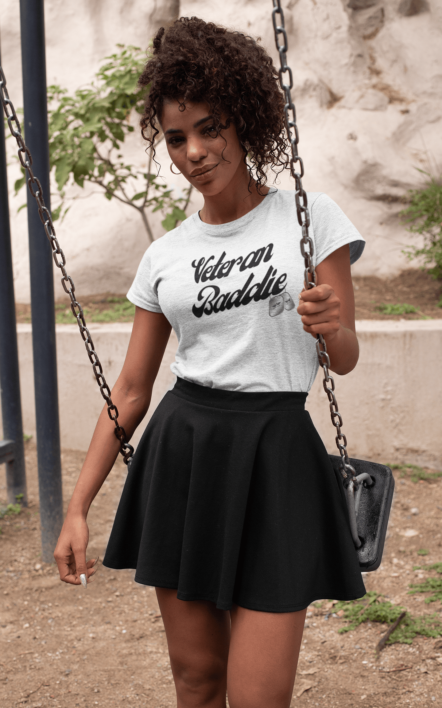 Young woman with curly hair wearing a pink t-shirt with white text, denim shorts, and white sandals, standing smiling against a white background.