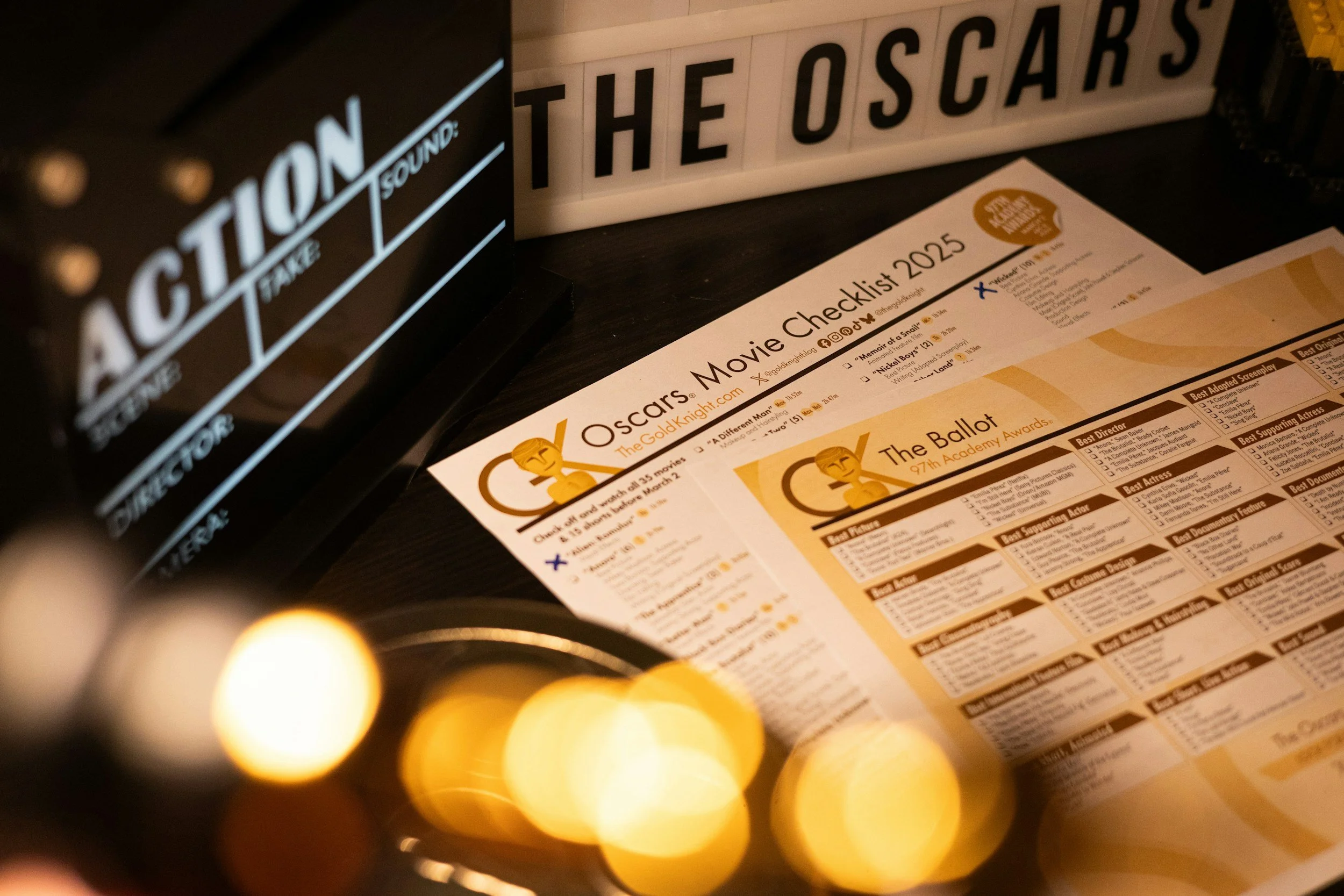 Close-up of a movie checklist and Oscar award information on a paper, with a blurred string of glowing yellow orbs in the foreground, and illuminated signage spelling 'THE OSCARS' in the background.