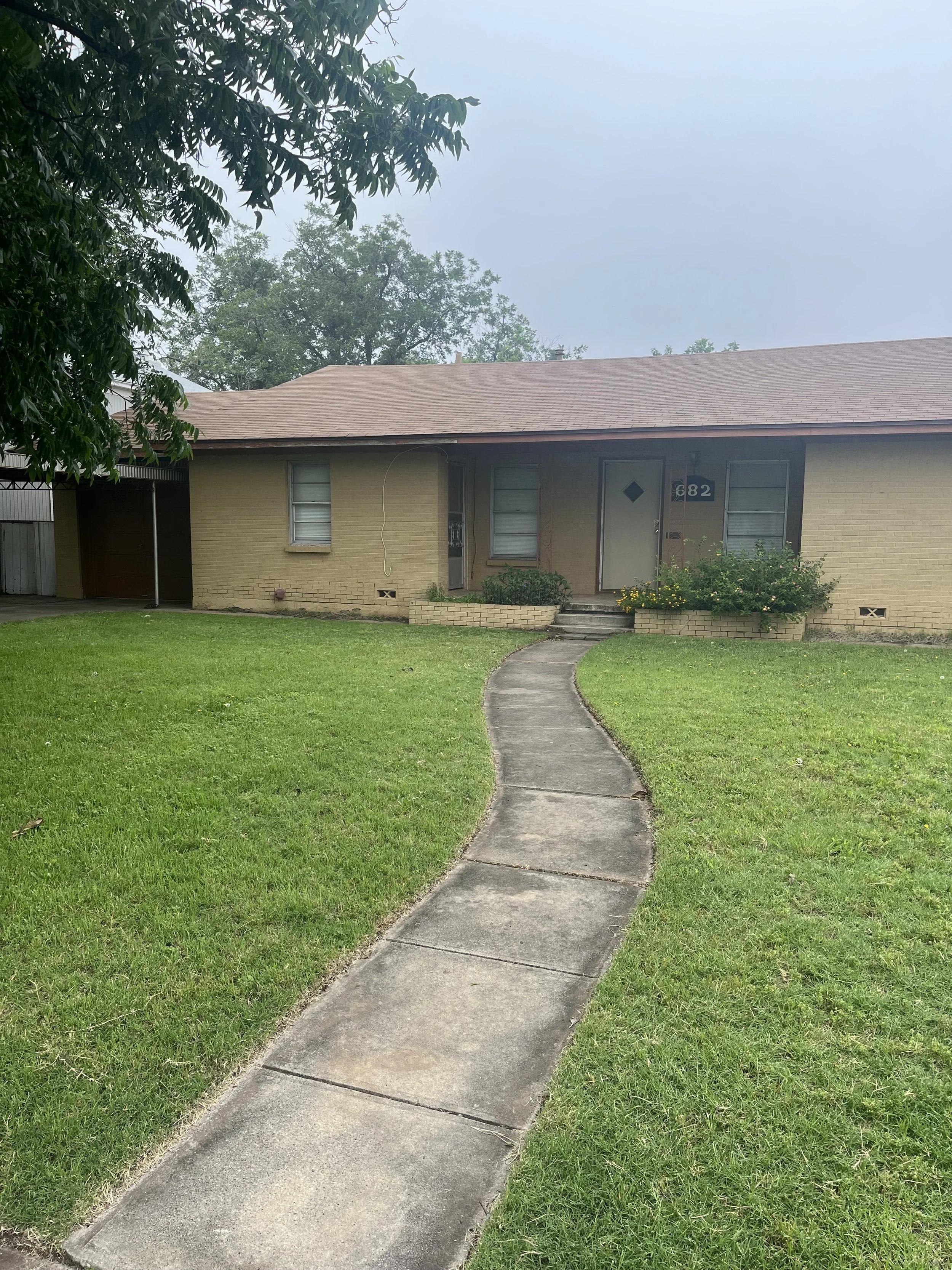 Front yard of a house with a curved sidewalk leading to the front door, a lawn, bushes, and trees in the background under a cloudy sky.