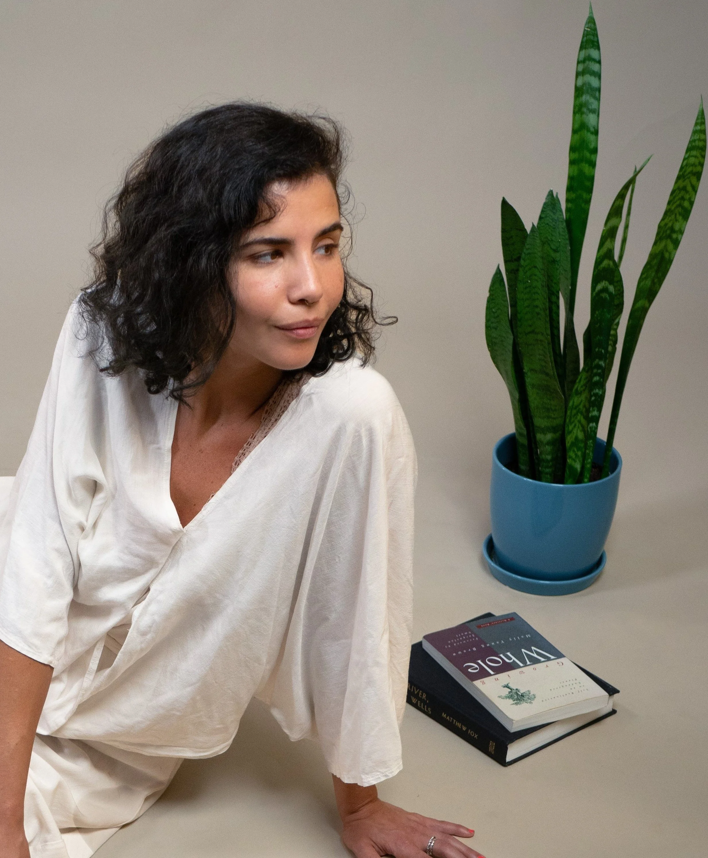 A woman with curly black hair in a white dress sitting on the floor next to a potted snake plant and two books.