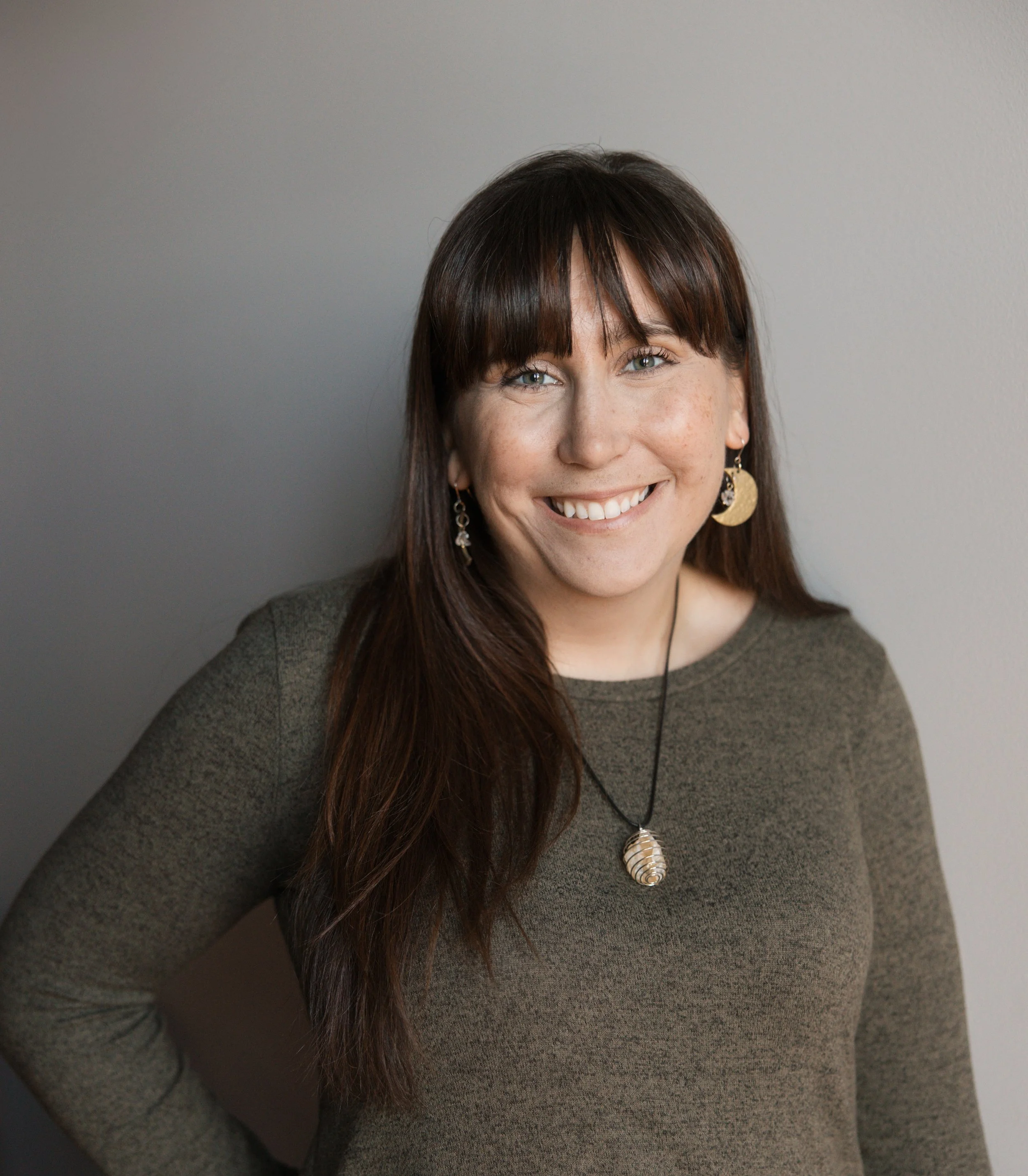 Katelyn Thoresen, a brown haired woman wearing a medium brown sweater, a shell on a necklace, and moon shaped earrings.
