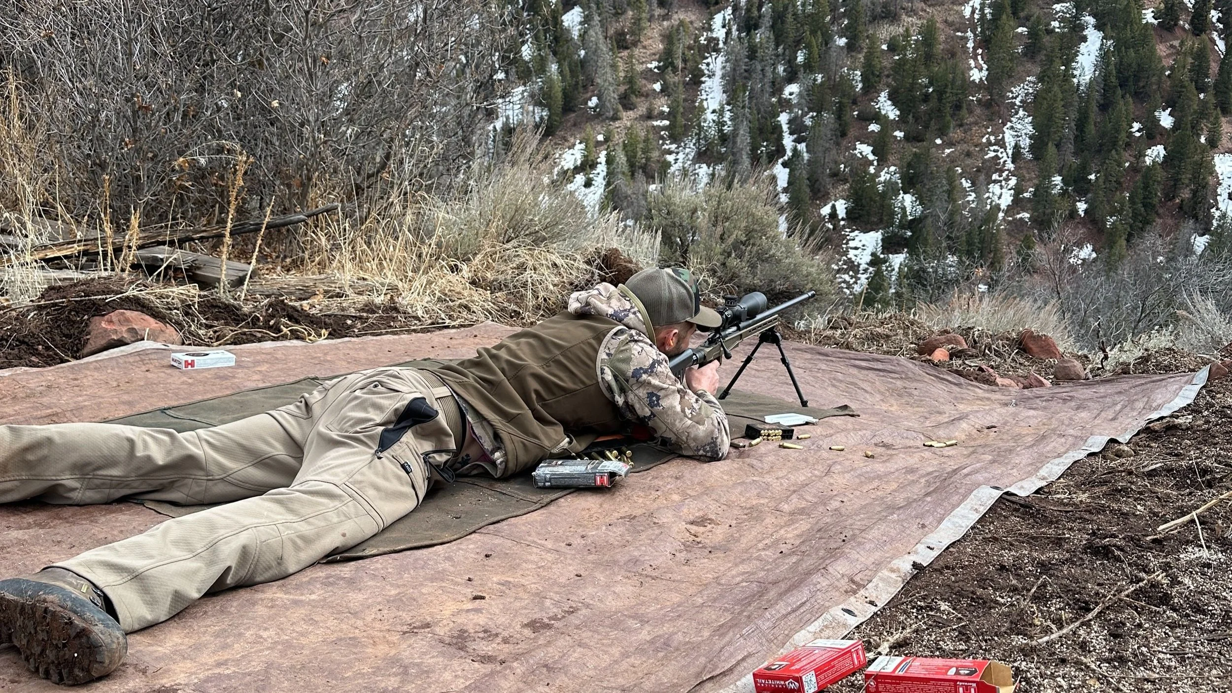 A person lying prone on a tarp in a mountainous outdoor area, aiming through a sniper rifle with a scope, surrounded by ammunition and equipment. Long-distance shooting lessons. 1,000 yard shooting.