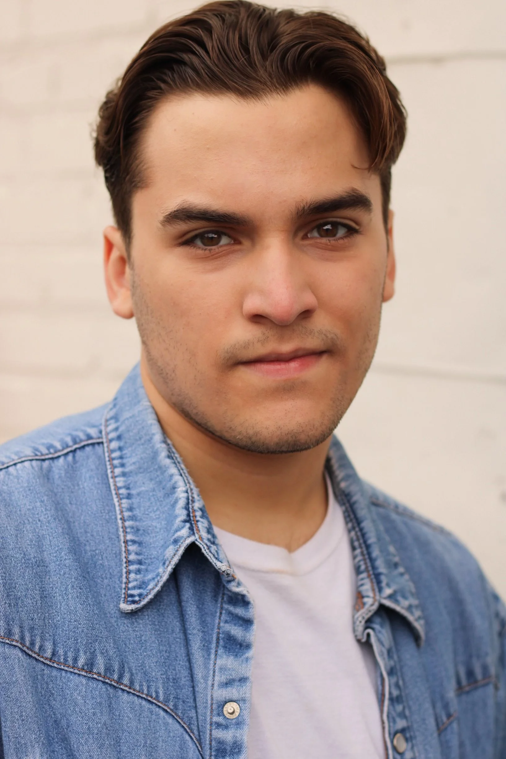 Close-up portrait of a young man with brown hair, wearing a blue denim jacket over a white t-shirt, standing against a light-colored brick wall.