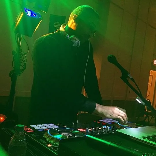 A DJ performing at a club with green lighting, wearing headphones and operating DJ equipment with a microphone nearby.