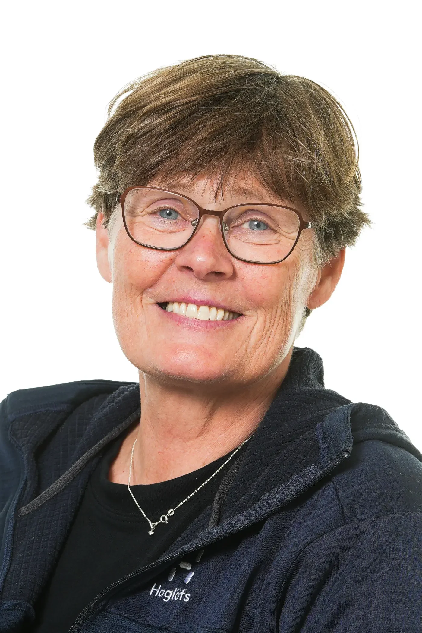 Et close-up af en glade ældre kvinde med brunt, skåret hår, briller og smil, iført en sort t-shirt og en mørk jakke, mod en hvid baggrund.
