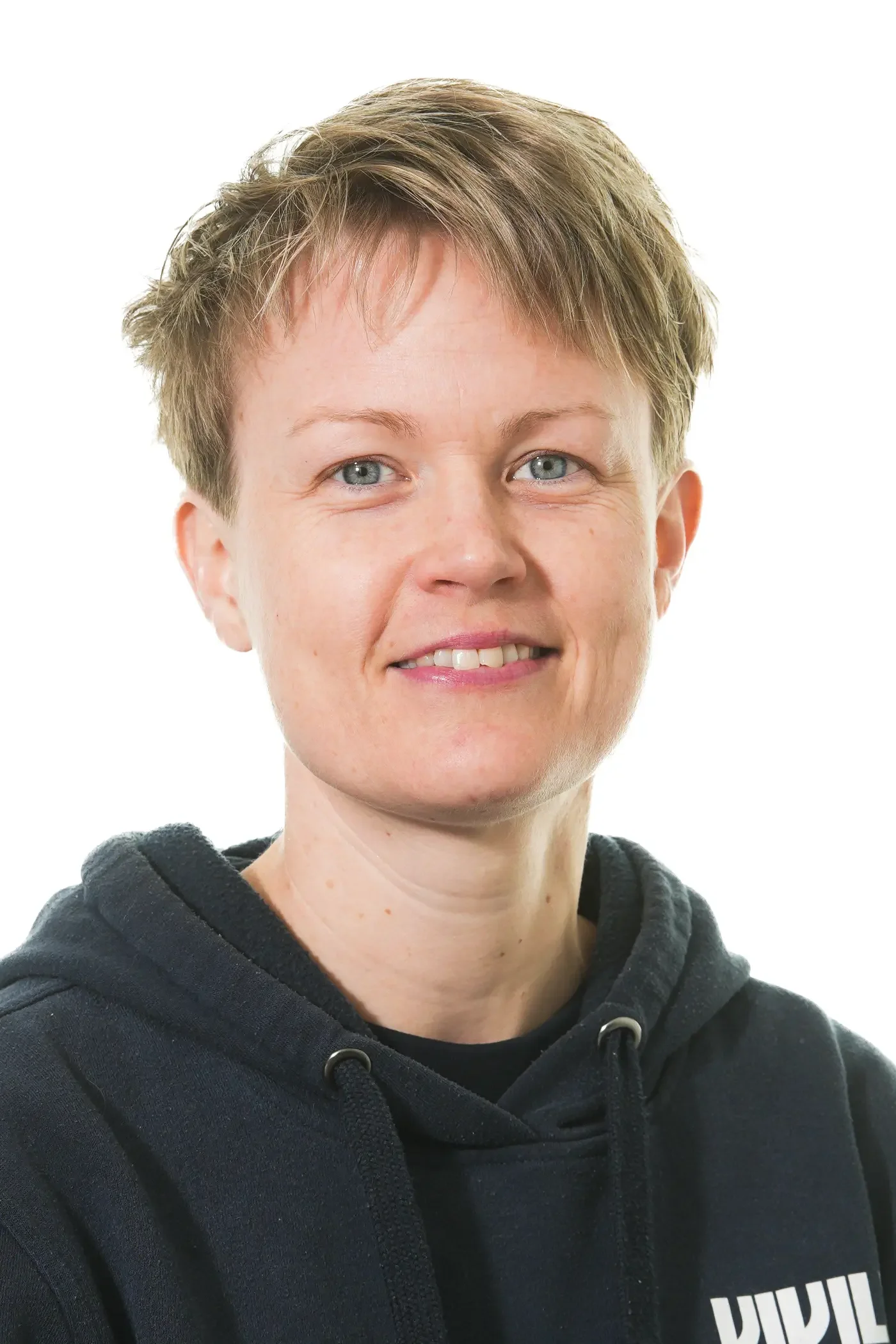 Billedet viser en kvinde med kort, blond hår, blå øjne og et smil, der viser hendes tænder, iført en sort top.