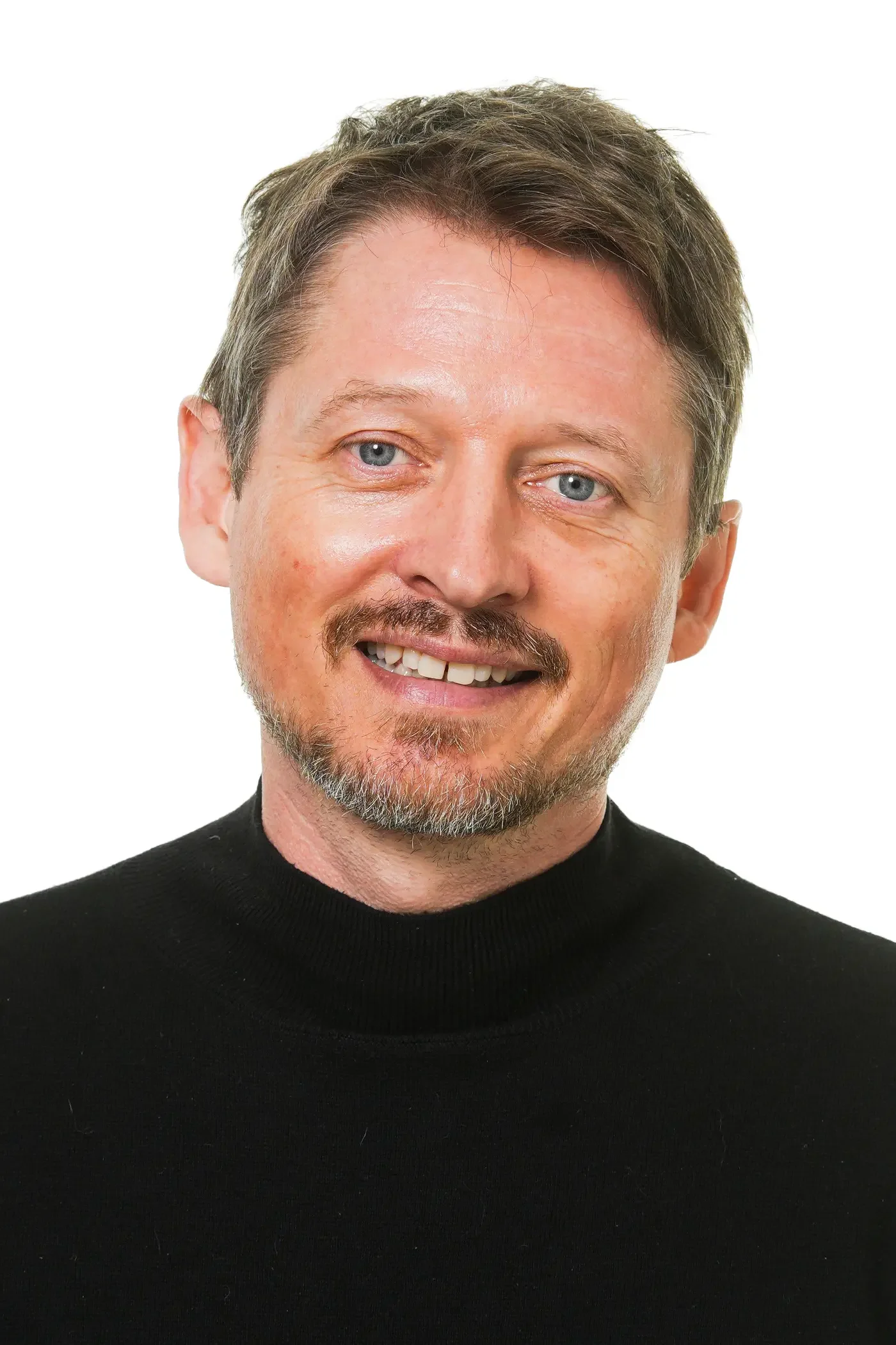 Et nærbillede af en glad mand med kort, brunt hår og blå øjne, iført en blå skjorte og en sort cardigan, mod en hvid baggrund.